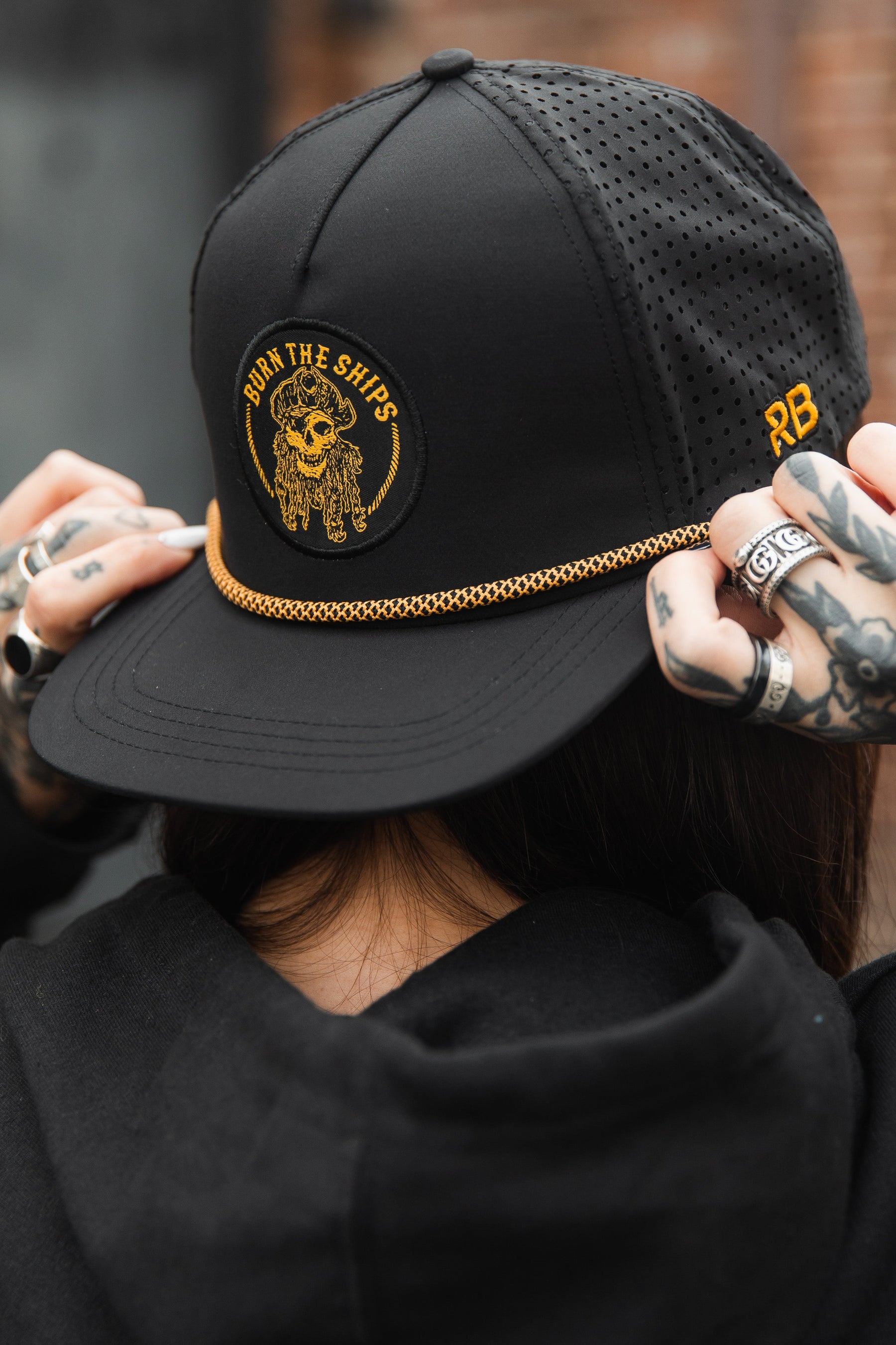 A person wears the Burn The Ships BLACKOUT Snapback by Relentless Betrayal, featuring gold embroidery of a skull and floral design with BURN THE SHIPS text. They have hand tattoos and a black hoodie. This snapback is one of the finest handmade hats in their collection.