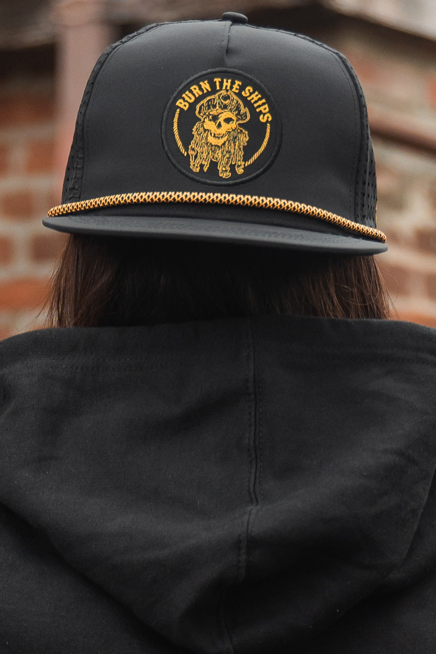 A person wears a Relentless Betrayal Burn The Ships BLACKOUT snapback, showcasing a yellow pirate skull with matching text. Theyre in a black hoodie, standing against an artfully blurred brick wall that complements the handmade hats unique style.