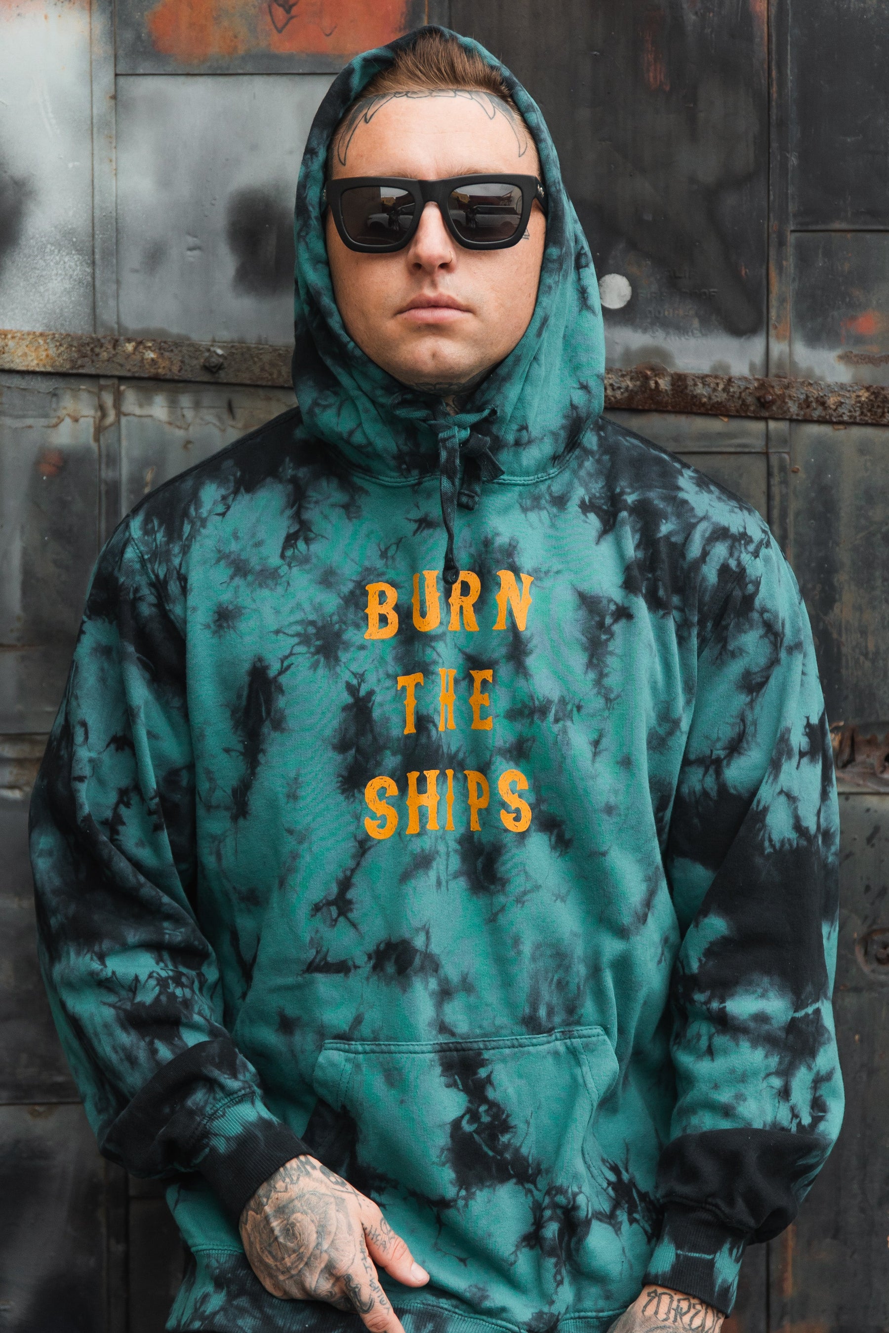 A person stands against a metal wall wearing the Burn The Ships Tie Dye Hoodie from Relentless Betrayal. The teal and black unisex hoodie boldly features BURN THE SHIPS in yellow. Paired with black sunglasses and tattoos, it embodies edgy style and comfort in its cotton polyester blend.