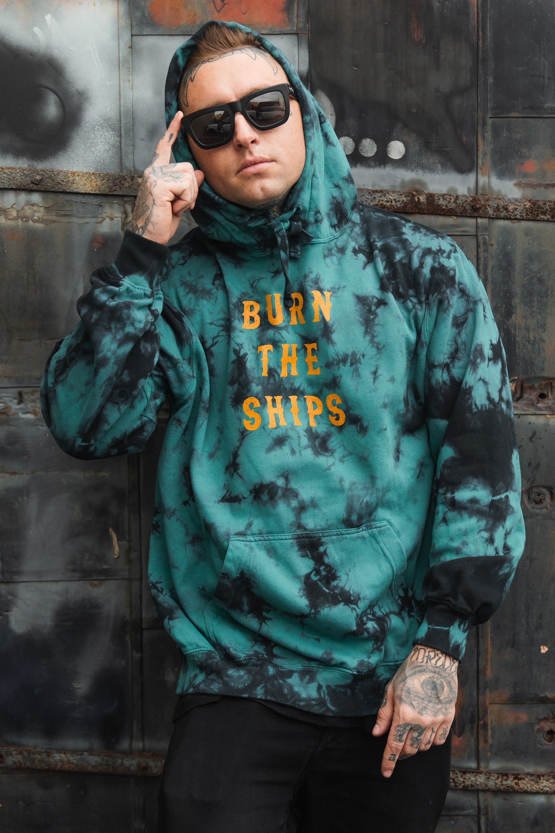 A person wears a Relentless Betrayal Burn The Ships green and black tie-dye hoodie, crafted from a cozy cotton-polyester blend. Posing against a metal wall, they display tattoos on their hand and neck while adjusting their sunglasses.