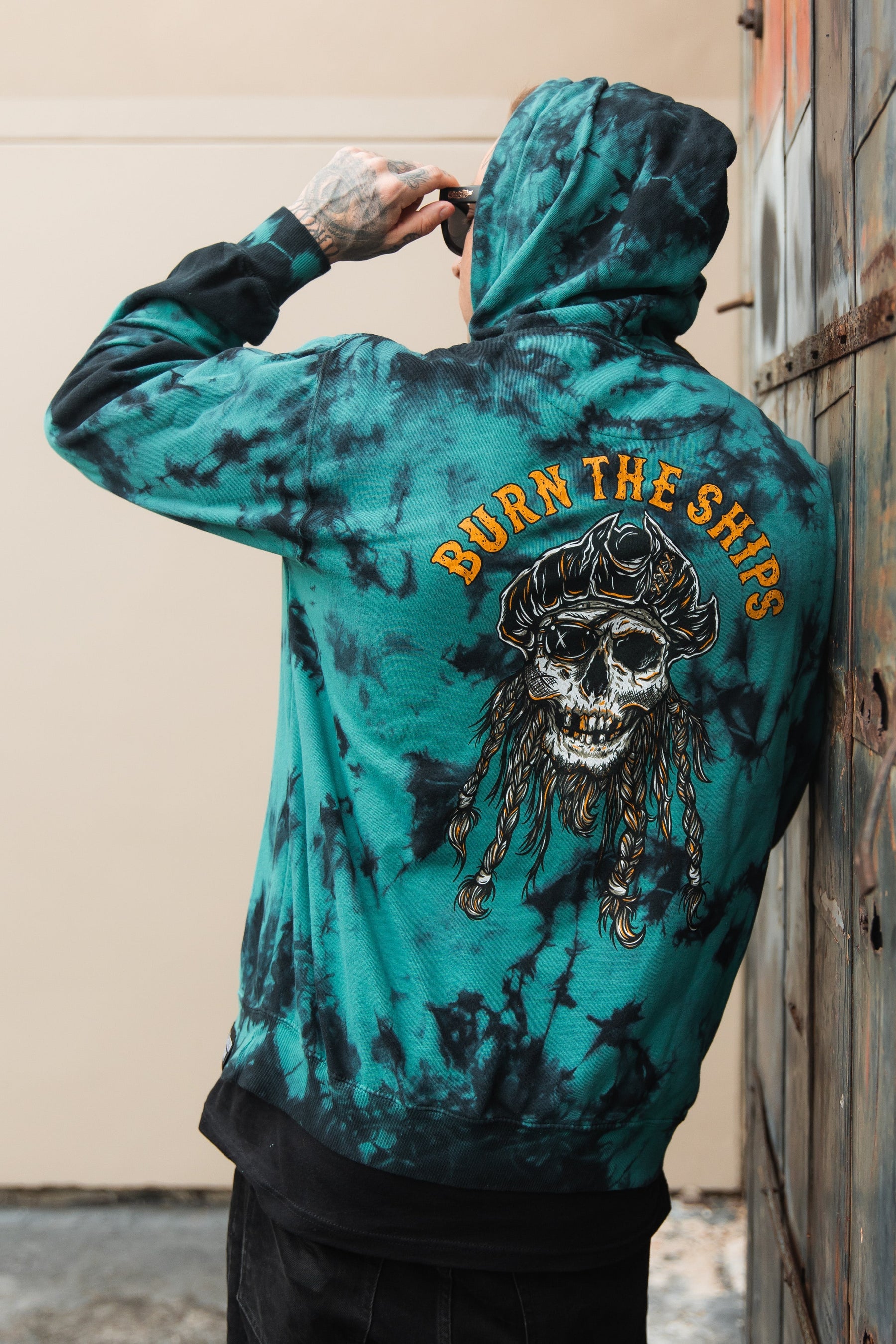 A person wearing the Relentless Betrayal Burn The Ships Tie Dye Hoodie with a pirate skull design adjusts their sunglasses while standing beside a weathered wall. The unisex hoodie, in blue tie-dye, features the phrase BURN THE SHIPS and is made from a cozy cotton-polyester blend.