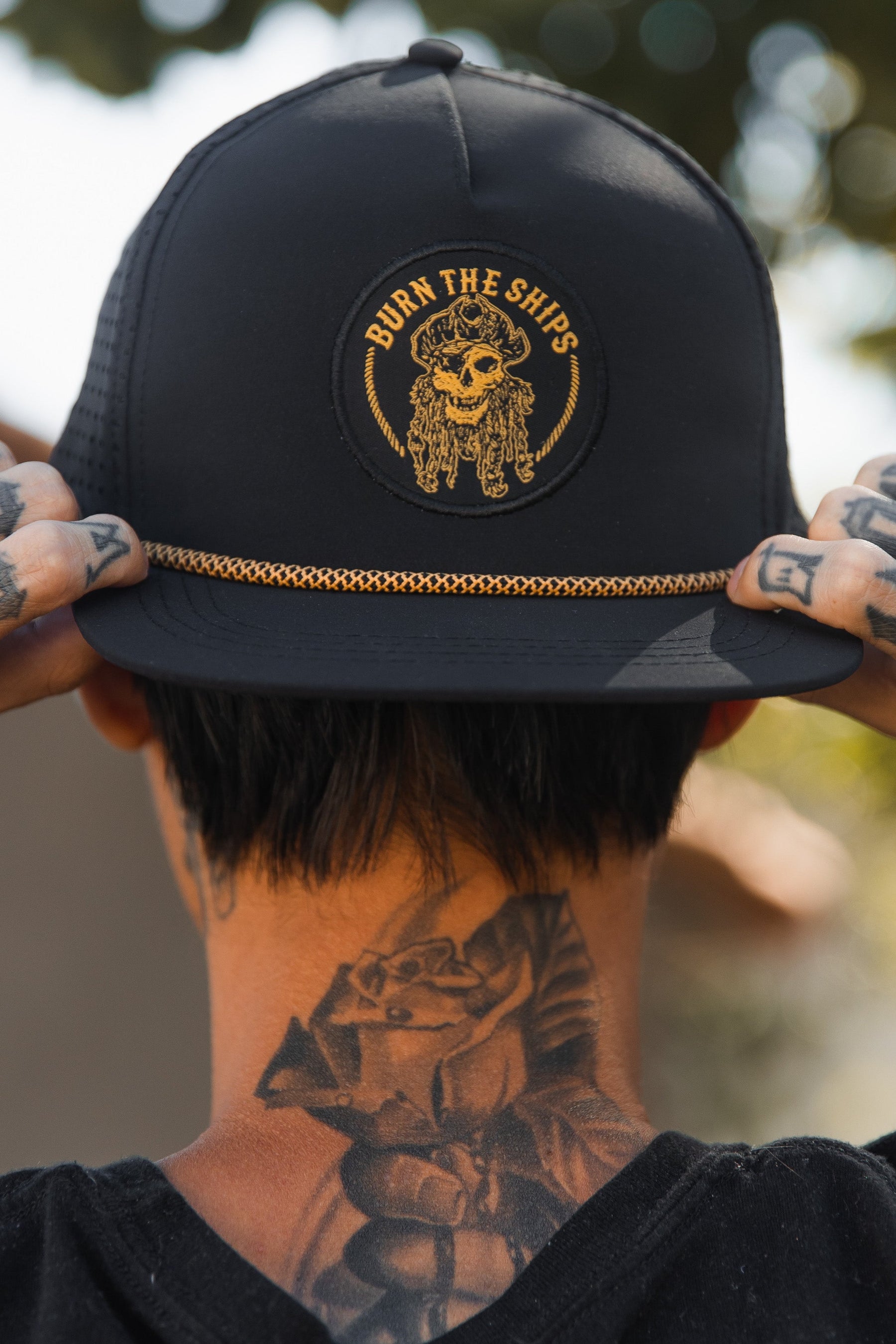 A tattooed person wearing the Relentless Betrayal Burn The Ships BLACKOUT Snapback, featuring a gold pirate emblem and BURN THE SHIPS text, stands against a sunlit, tree-blurred background.