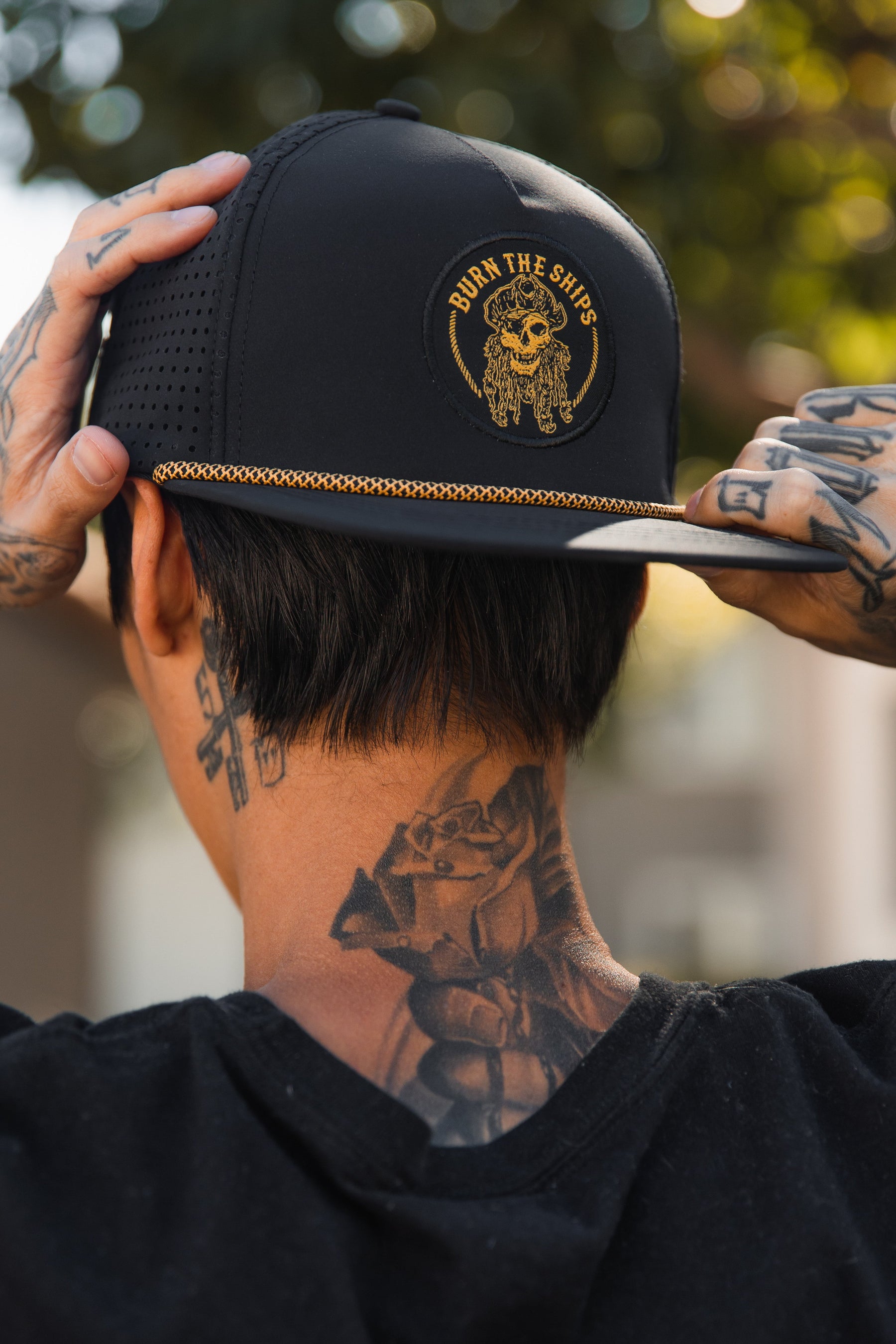 An individual with tattooed arms and neck, their back to the camera, adjusts a Burn The Ships BLACKOUT Snapback by Relentless Betrayal. The cap displays a gold embroidered skull and ships wheel with BURN THE SHIPS boldly emblazoned. A blurred background adds mystery.