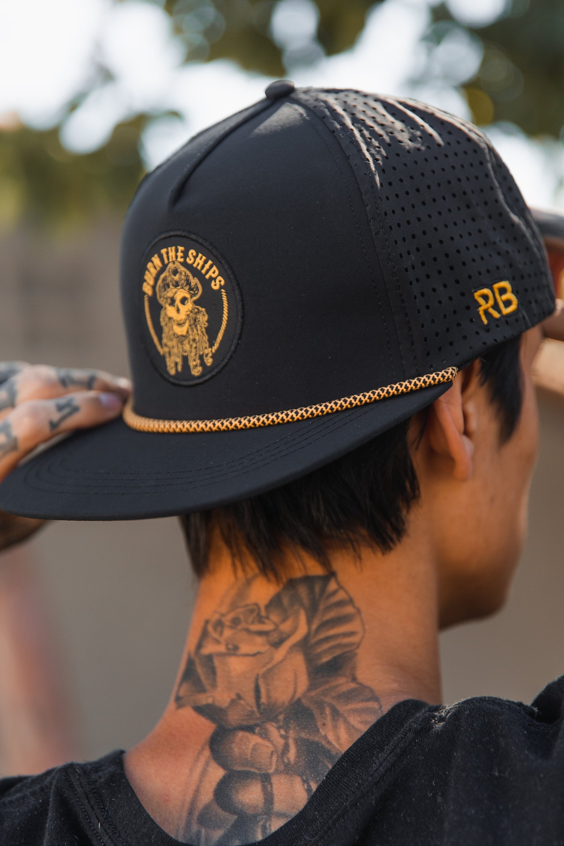 Wearing the Burn The Ships BLACKOUT Snapback from Relentless Betrayal, featuring a sailors emblem and RB on the side, a tattooed individual showcases inked arms with a rose tattoo against a softly blurred tree background.
