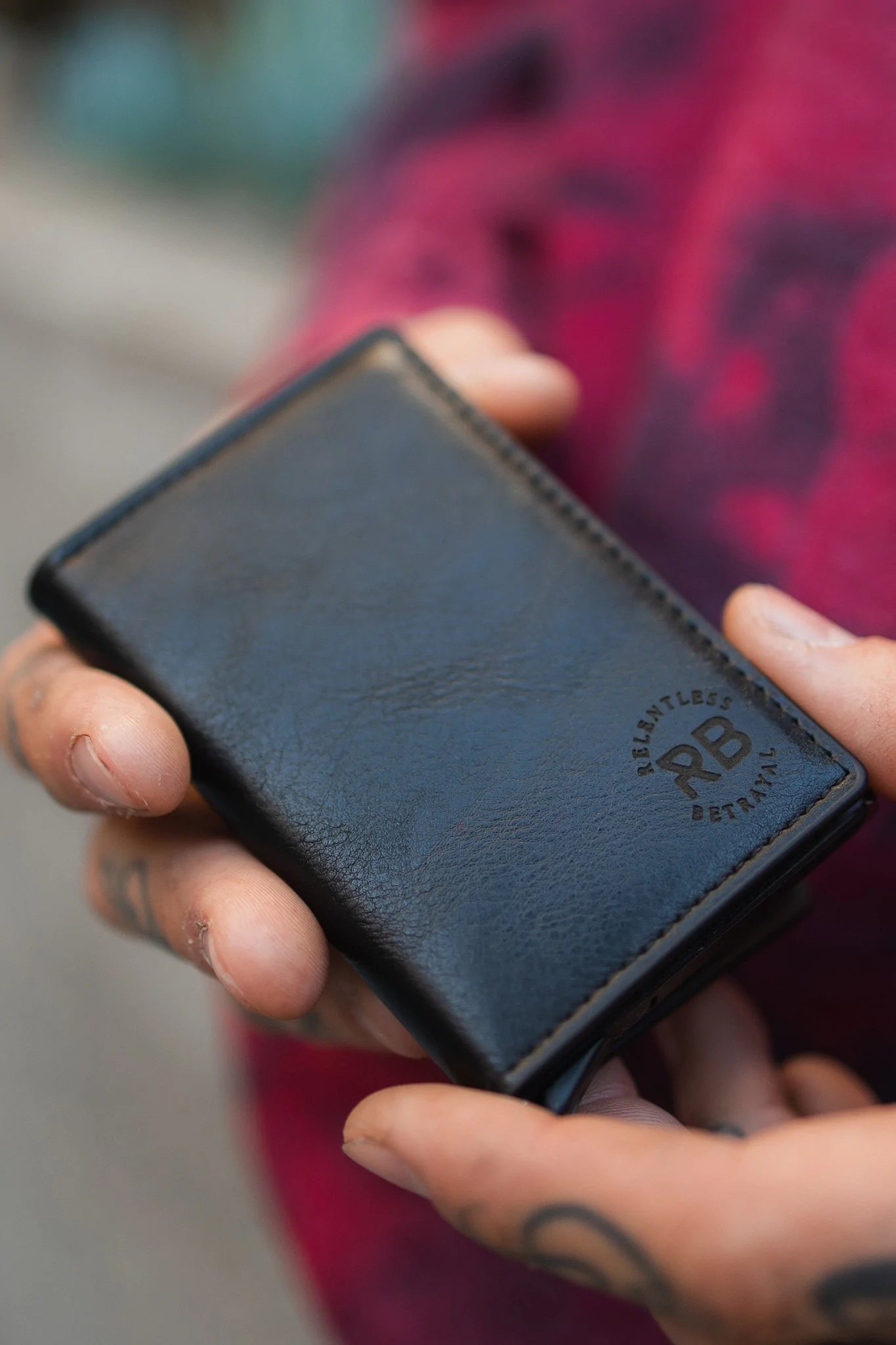 A person with finger tattoos holds a Relentless Betrayal Wallet by Relentless Betrayal, made from synthetic leather and embossed with RELENTLESS DETAILS, against their textured reddish outfit. The background is artistically blurred.