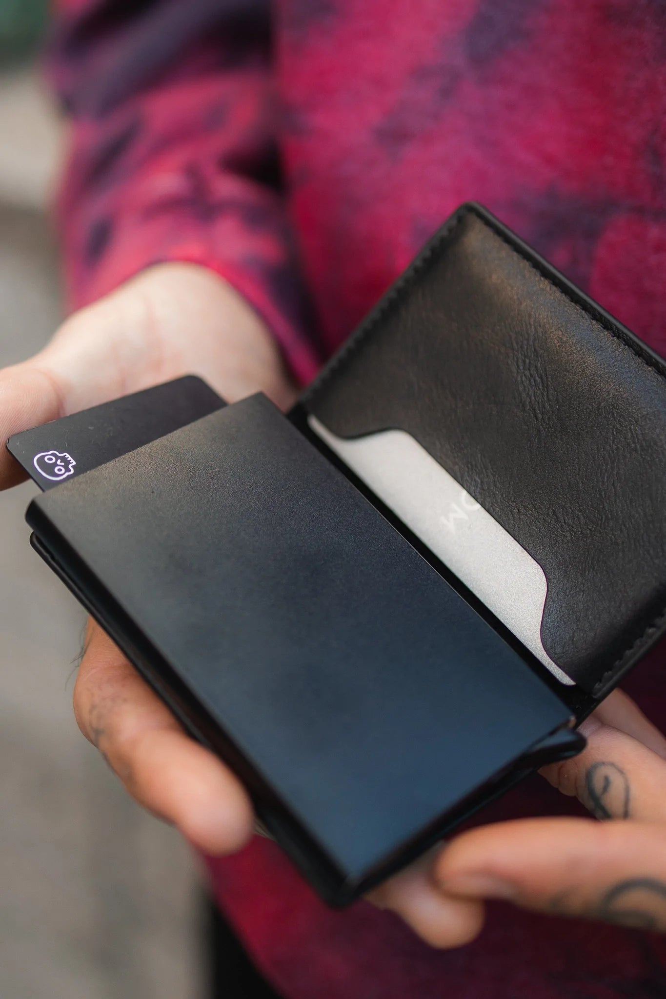 A person with a Relentless Betrayal synthetic leather wallet, showcasing an exposed card marked 6+, has tattooed fingers that complement their red and purple patterned shirt.