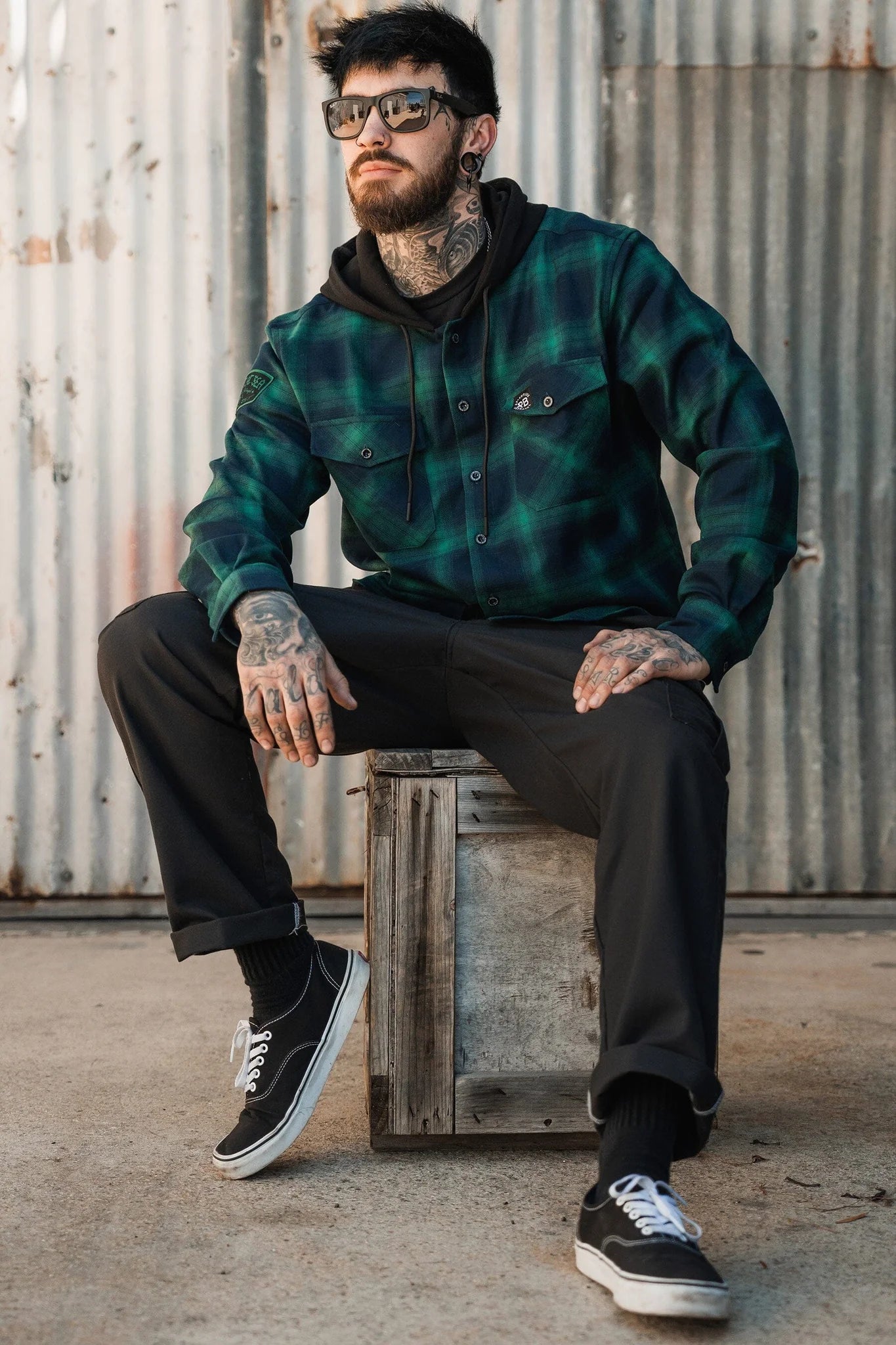 A man with sunglasses and tattoos sits on a wooden box in an industrial setting, wearing Relentless Betrayals Lost At Sea Premium Hooded Flannel in green plaid over a black hoodie. He pairs it with dark pants and black sneakers, framed by corrugated metal that echoes his rugged style.