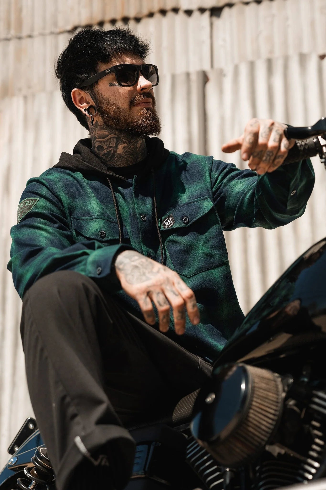A tattooed man, in sunglasses and a Lost At Sea Premium Hooded Flannel by Relentless Betrayal, sits on a motorcycle. The ultra-soft flannel adds contrast to his edgy vibe as he gazes thoughtfully against a corrugated metal backdrop.
