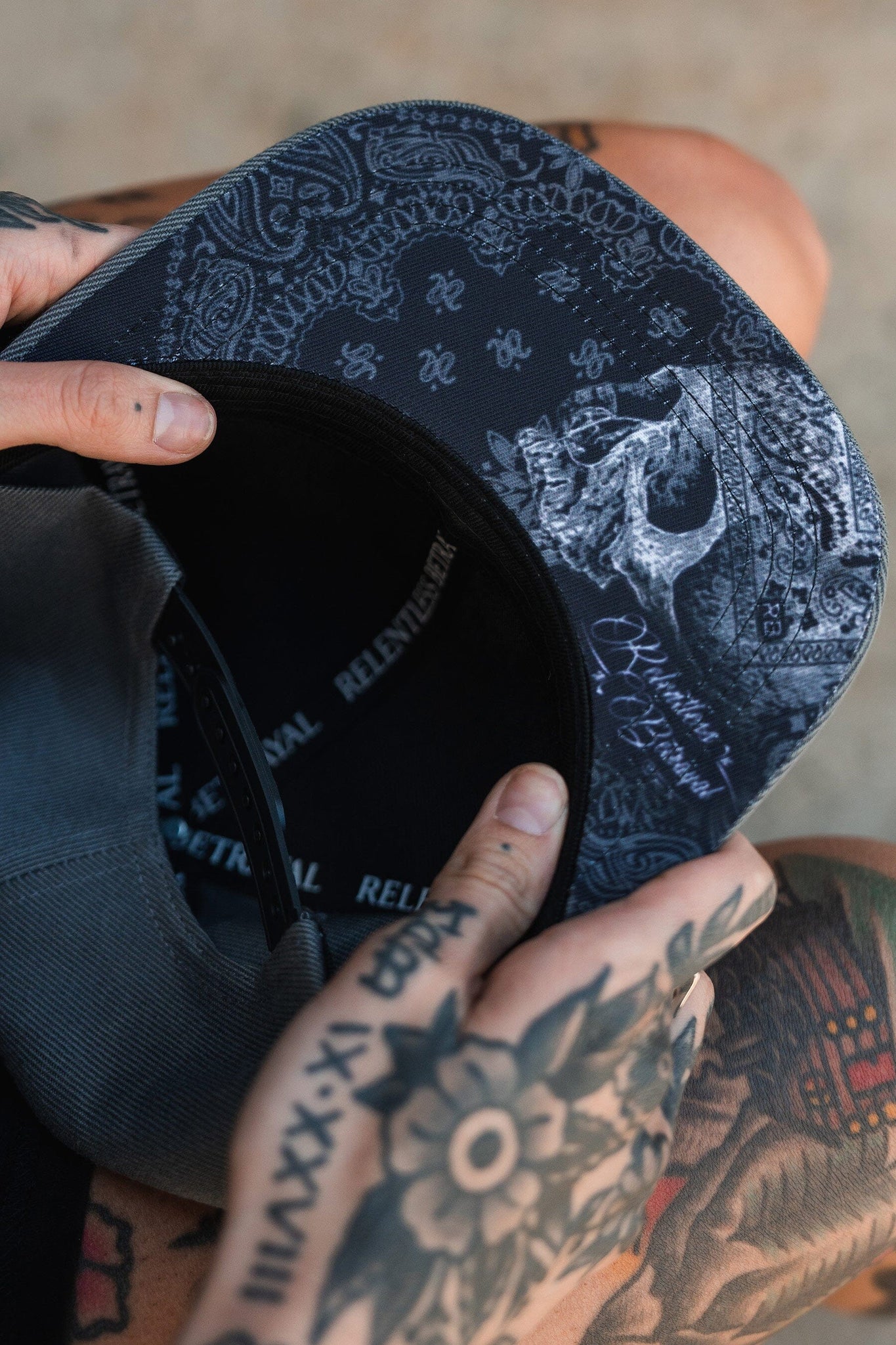 Close-up of tattooed hands holding the Skilled Sailor Snapback by Relentless Betrayal, highlighting its detailed black and gray skull design with intricate patterns. The person wears dark clothing that perfectly complements the unique style of these handmade hats.
