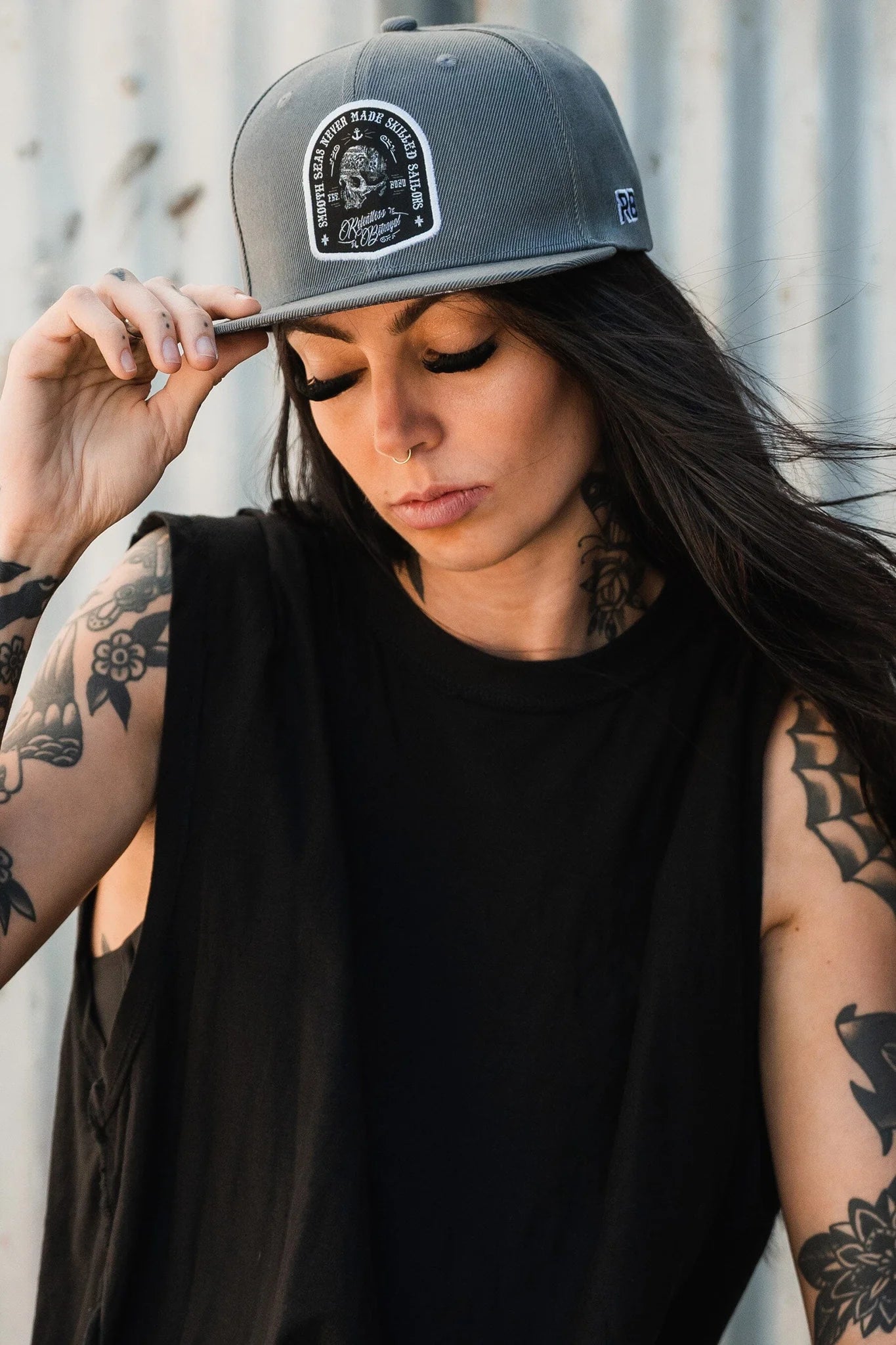 The woman with long dark hair and tattooed arms wears a Skilled Sailor Snapback by Relentless Betrayal and a sleeveless black top, standing against a corrugated metal background. She holds the hats brim thoughtfully, lost in contemplation.