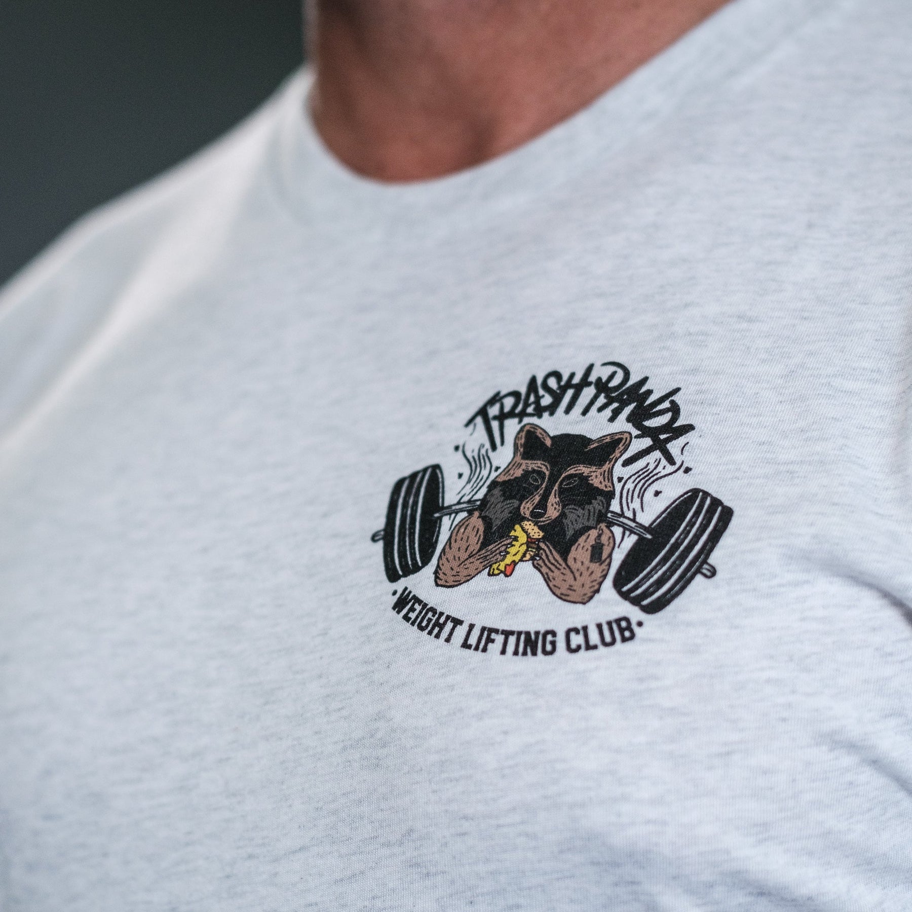 A close-up showcases a person in the Tango Charlie Apparels light gray tri-blend Trash Panda Weight Lifting Club Tee featuring a playful raccoon lifting weights logo encircled by the club’s name, combining strength with style.