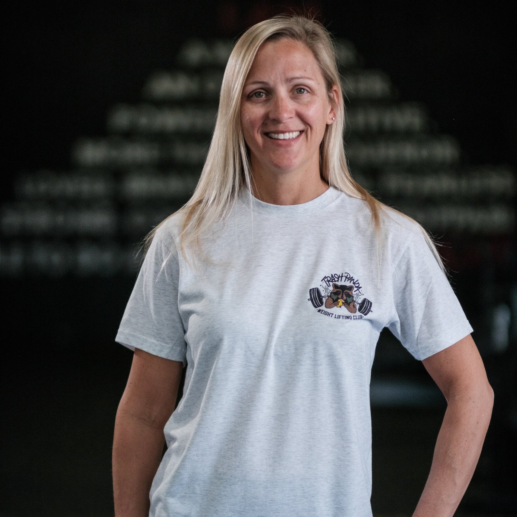 A smiling person with long blonde hair wears a Tango Charlie Apparel Trash Panda Weight Lifting Club Tee, light gray and tri-blend, against a dark, blurred background highlighting a pyramid of weights for an energetic vibe.
