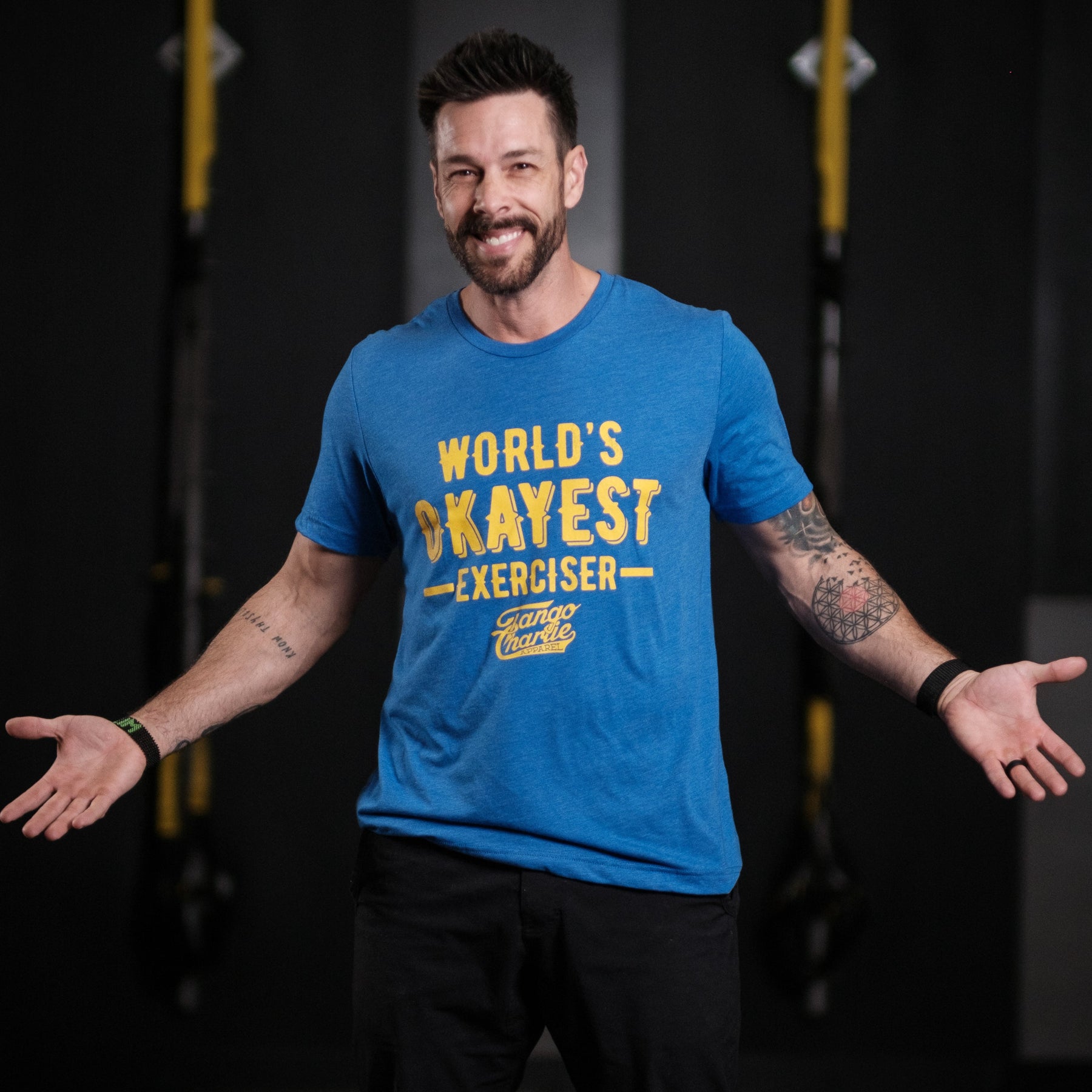 A bearded man stands smiling with arms outstretched, wearing a comfy Worlds OKAYEST Exerciser Tee by Tango Charlie Apparel. The cotton/poly blend shirt features bold yellow text against a blurred gym backdrop, creating an amusing scene.