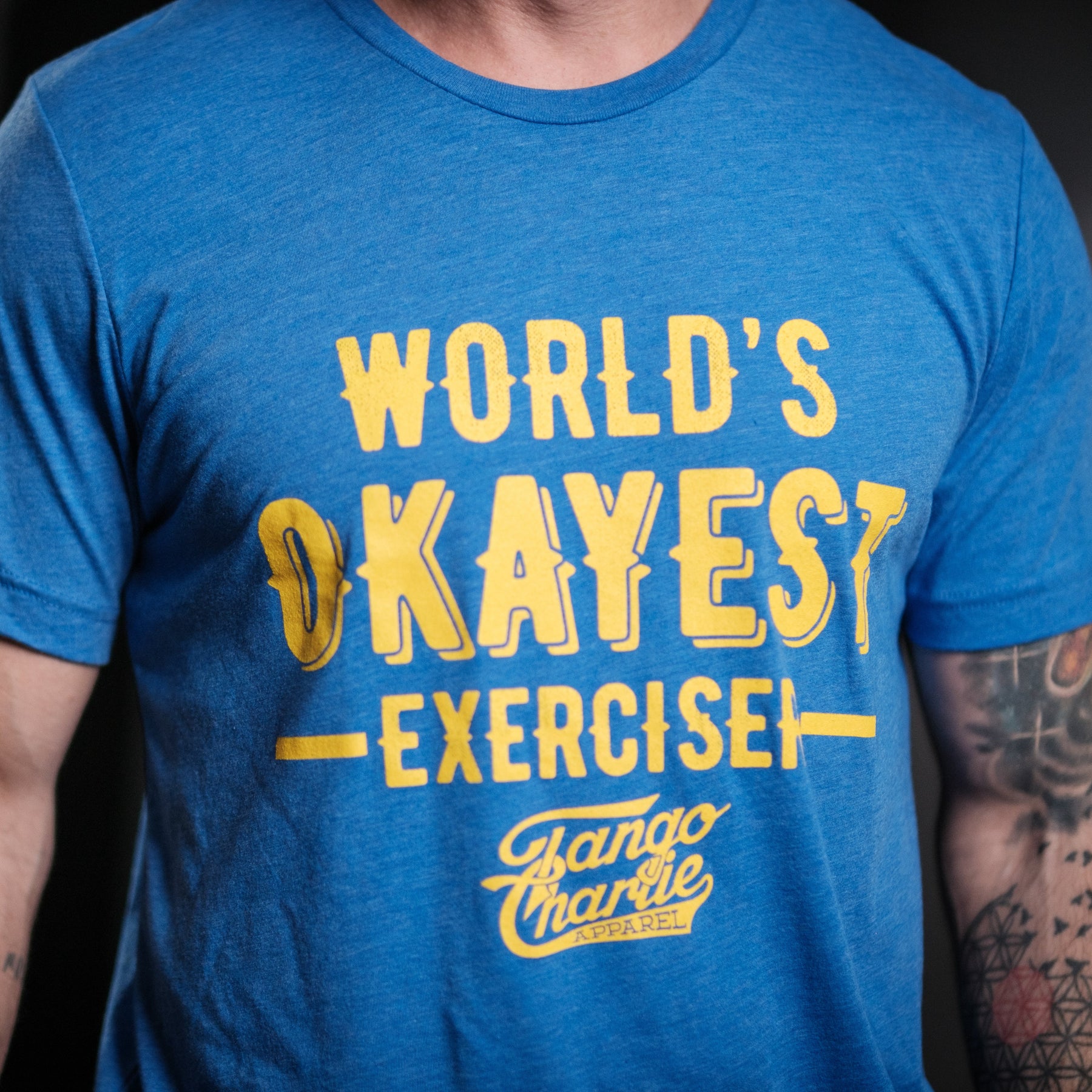 Wearing a comfy cotton/polyester blend tee, a person showcases the blue Worlds OKAYEST Exerciser T-shirt by Tango Charlie Apparel, complemented by tattoos on their right arm for an edgy look.