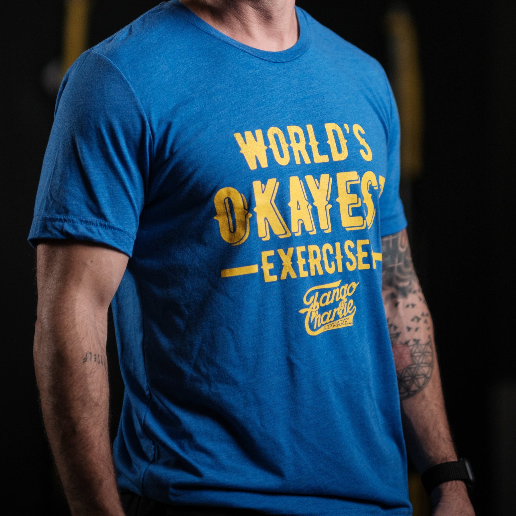 A person dons the Tango Charlie Apparels Worlds OKAYEST Exerciser Tee, a comfy blue cotton/poly blend with yellow text. Their tattooed arm adds flair against a dark, blurred background.