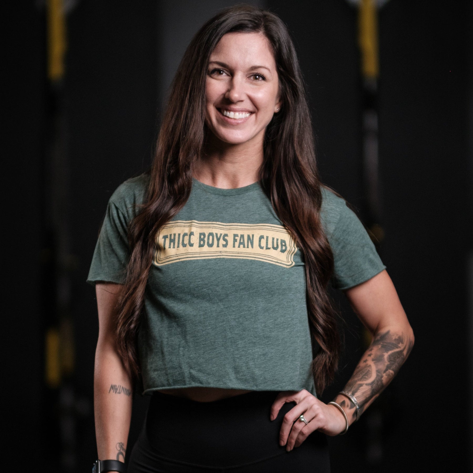A person with long, dark hair smiles confidently in a gym, wearing a Tango Charlie Apparel green THICC Boys Fan Club Crop T-Shirt, surrounded by the blurred outlines of exercise equipment.