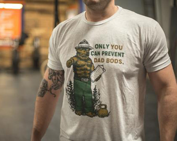 Wearing Tango Charlie Apparels Only You Can Prevent Dad Bod tri-blend T-shirt, a person showcases a whimsical bear in a ranger hat with clipboard. A tattoo graces their arm as they pose against a blurred indoor background.