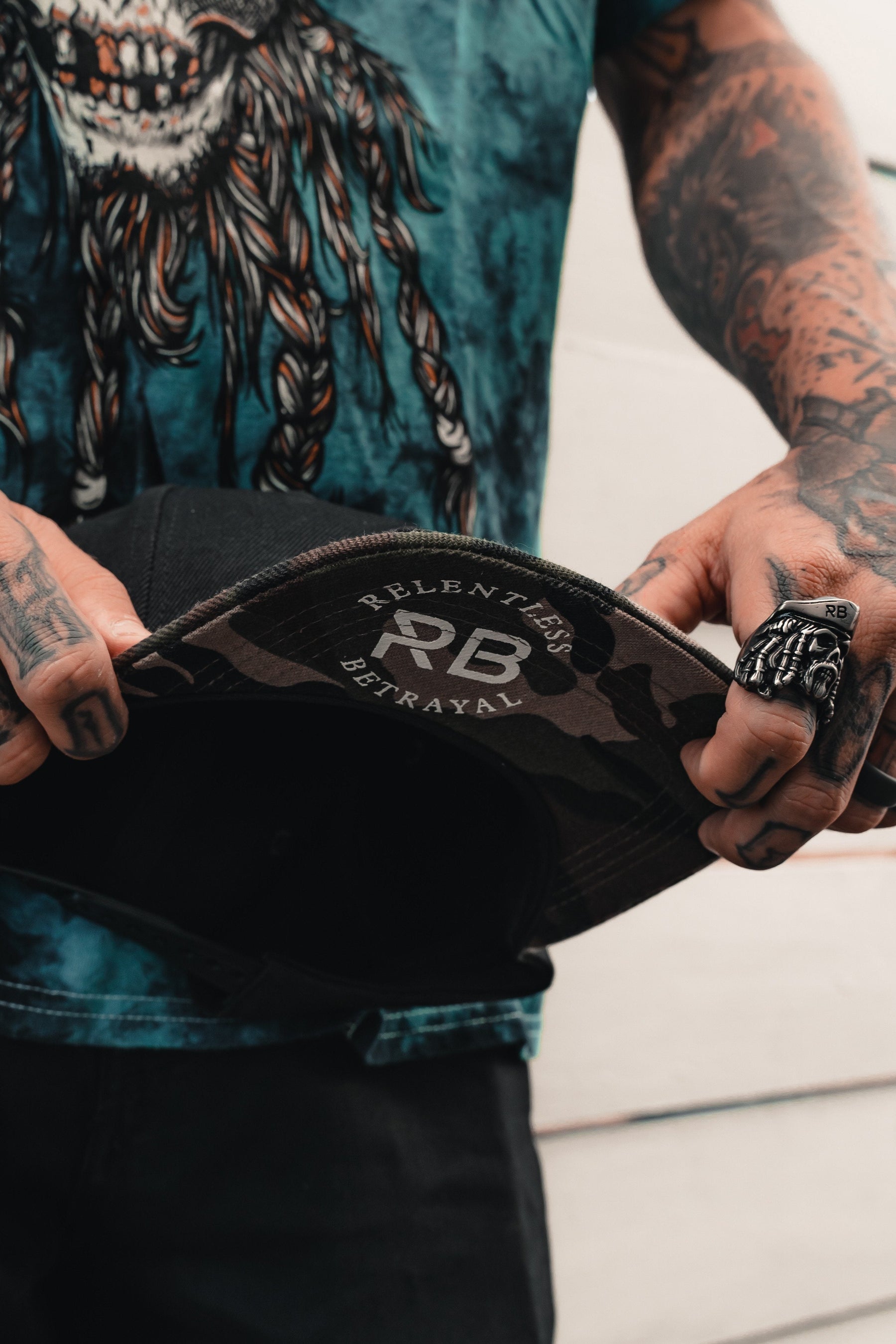 A person with tattooed arms holds the Burn The Ships Camo Snapback by Relentless Betrayal, RB Relentless Betrayal printed inside the brim. They sport a detailed ring and a colorful, graphic T-shirt, exuding an edgy vibe.