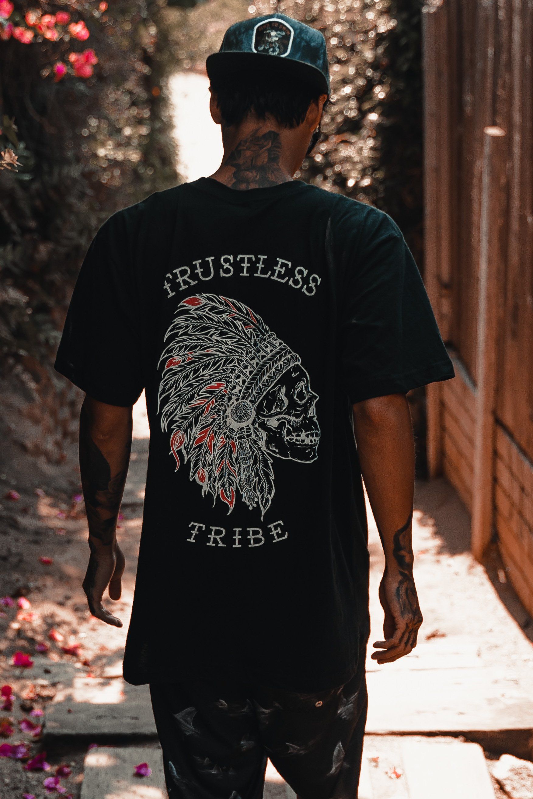 A person wearing the Trustless Chief Tee - Red by Relentless Betrayal strolls down a sunlit path. This shirt features a skull with a headdress and Trustless Tribe text. With a black cap and tattoos, they embody the essence of the trustless chief.