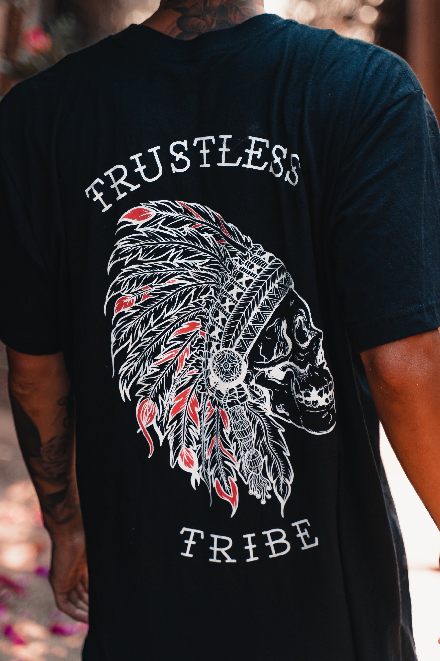A person wears the Trustless Chief Tee - Red by Relentless Betrayal, with a bold skull and feathered headdress graphic and Trustless Tribe text, epitomizing anarchist fashion, perfect for bold rebel leaders.