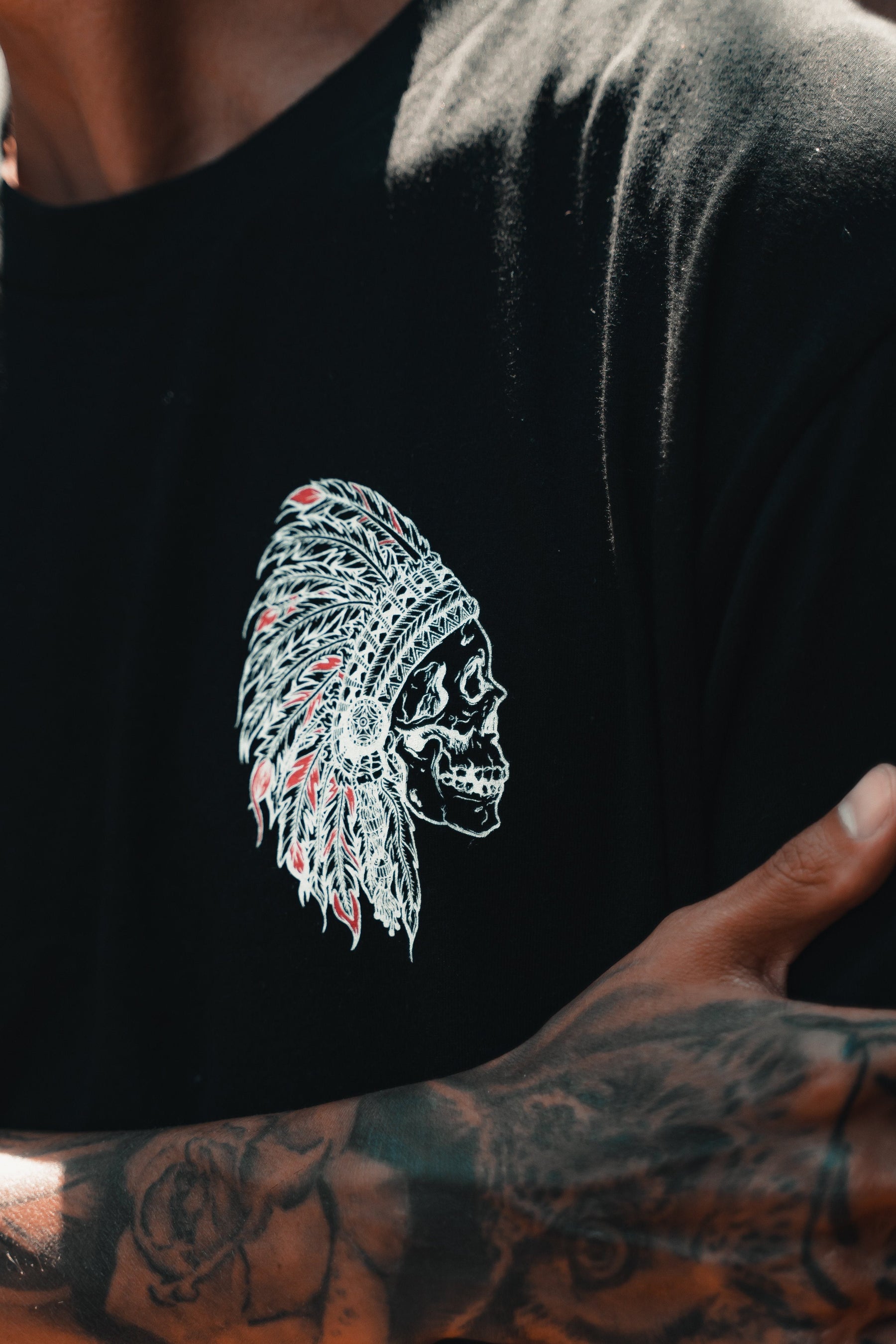 Close-up of a person wearing the Trustless Chief Tee - Red by Relentless Betrayal, featuring a skull with an intricate feather headdress. The anarchist vibe is enhanced by the persons tattooed arm crossed confidently in front of their body.