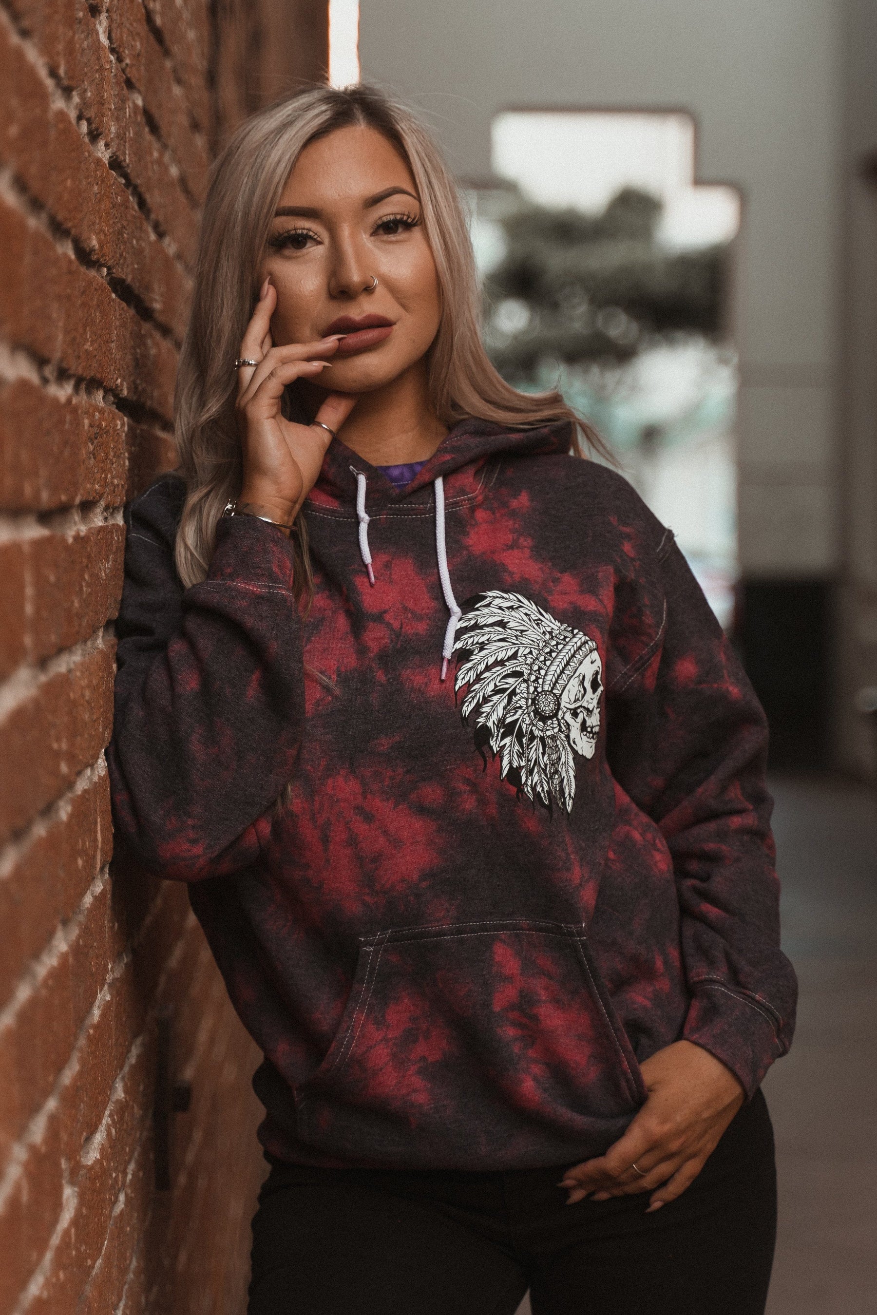 A person with long blonde hair leans against a brick wall, wearing a Relentless Betrayal Trustless Chief Red Tie Dye Hoodie featuring a Native American headdress graphic. One hand rests thoughtfully on their face, the other by their side.