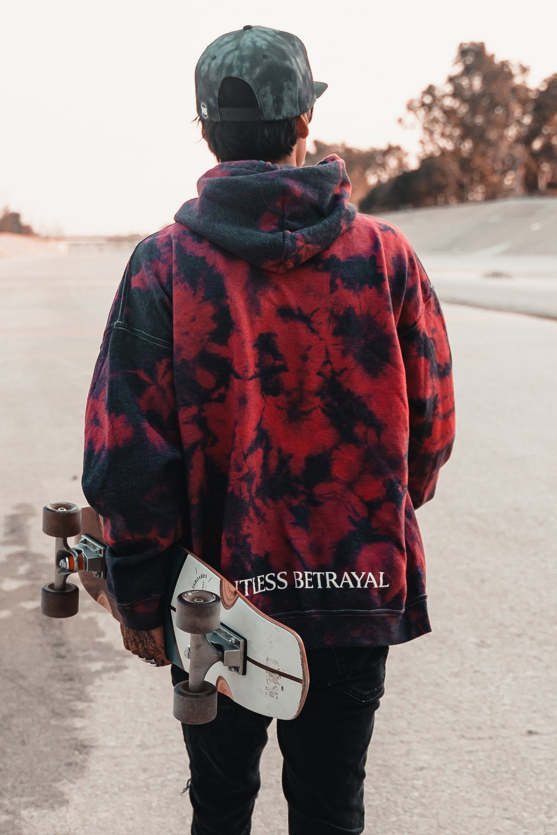 A person in a red tie-dye, cotton/polyester unisex Trustless Chief Hoodie by Relentless Betrayal and a cap stands on a paved path with a skateboard, trees lining the background.