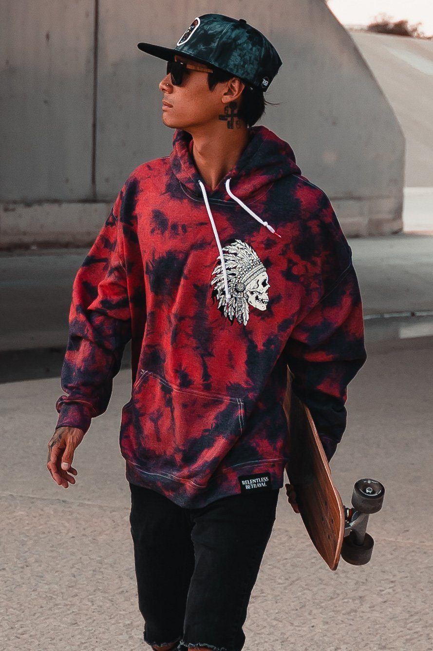 A young man strolls through the city, wearing a Trustless Chief Red Tie Dye Hoodie by Relentless Betrayal with a tribal skull design. He carries a skateboard, dons sunglasses and a cap, while his tattooed arm enhances his edgy vibe.