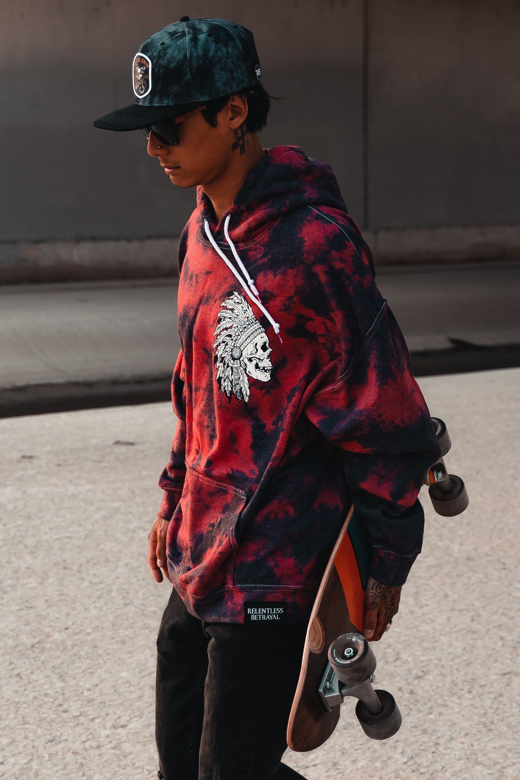 Wearing the Trustless Chief Red Tie Dye Hoodie by Relentless Betrayal, black pants, a dark cap, and sunglasses, a person strolls on a paved surface carrying a skateboard.