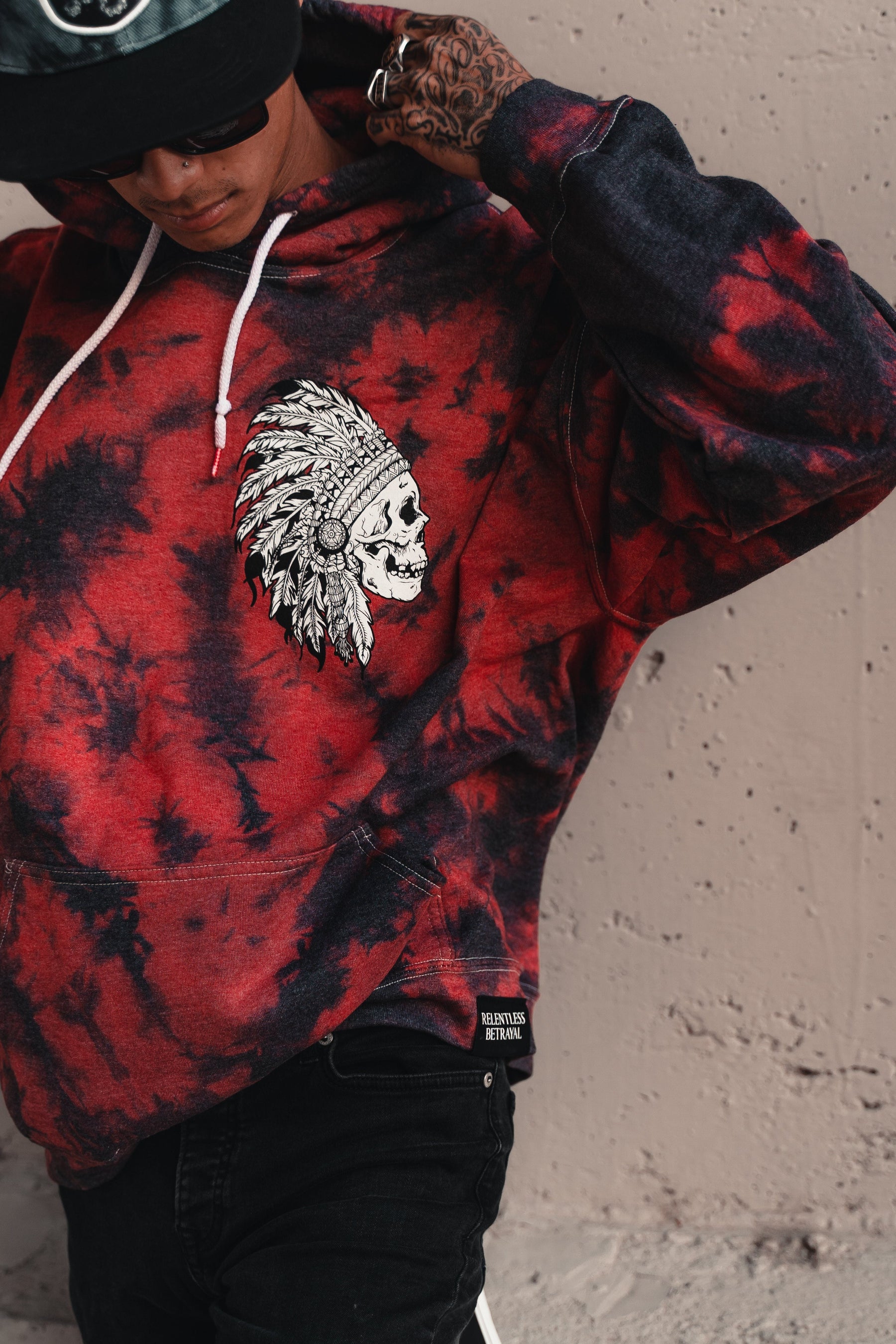 Donning the Trustless Chief Red Tie Dye Hoodie by Relentless Betrayal, featuring a skull and feather headdress design, a person adjusts their hoodie. Tattoos peek out as they sport sunglasses and a cap, blending edgy style with comfort in this unisex cotton/polyester hoodie.