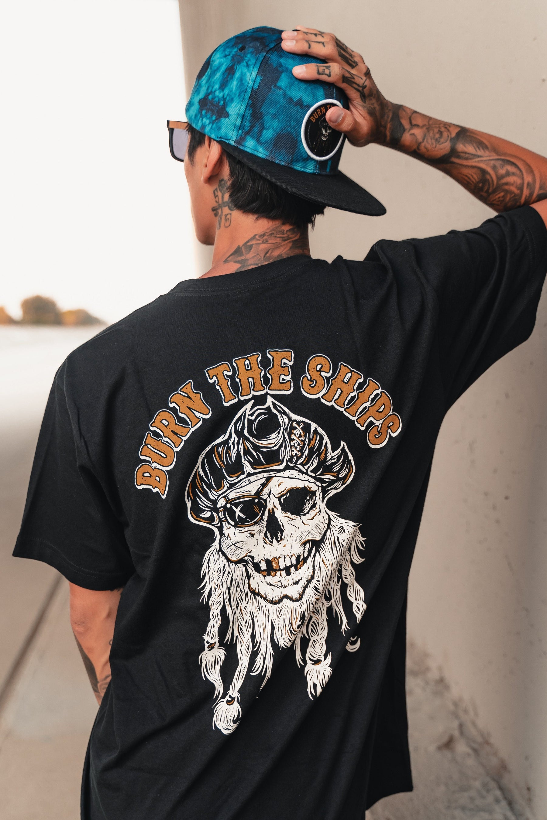 A tattooed individual stands outdoors, leaning against a wall and wearing a backwards blue cap and a Relentless Betrayal black cotton tee featuring BURN THE SHIPS with a pirate skull, hat, and beard.
