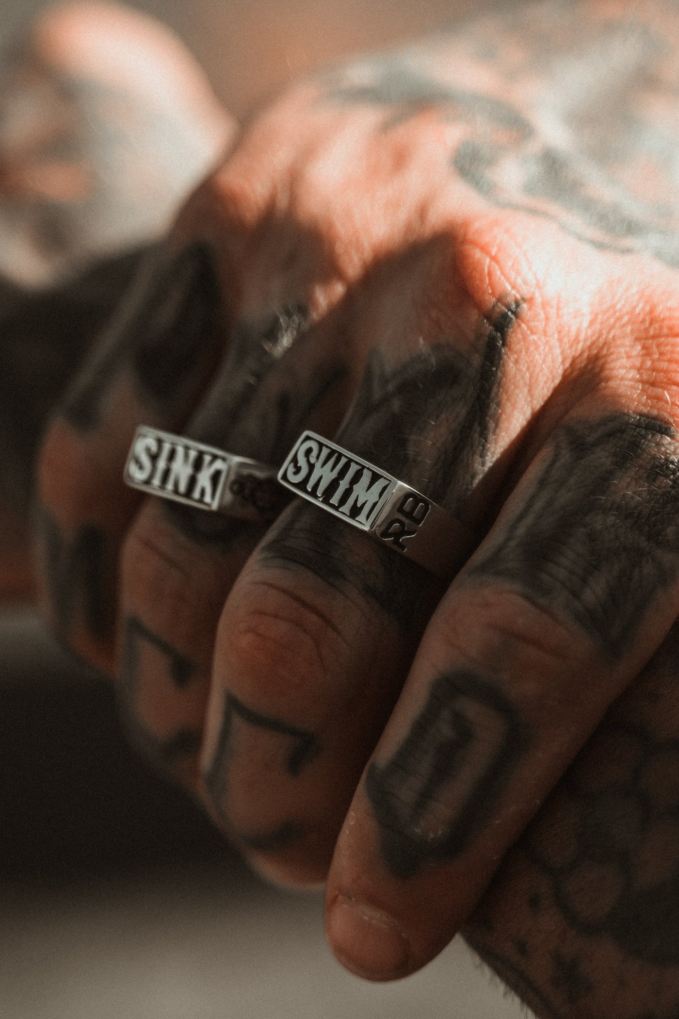 A tattooed hand adorned with Relentless Betrayals handcrafted Sink or Swim Rings, engraved with SINK and SWIM, basks in warm sunlight, showcasing the intricate details designed to complement both the tattoos and rings.