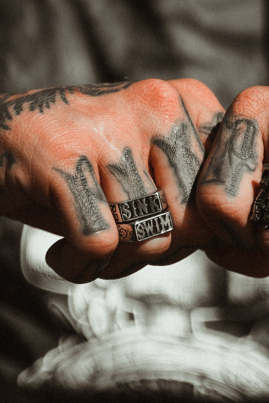 Close-up of tattooed hands adorned with Relentless Betrayals Sink or Swim Rings on the fingers. Crafted from stainless steel with SINK and SWIM engravings, these handcrafted rings paired with tattoos exude a bold, artistic style.