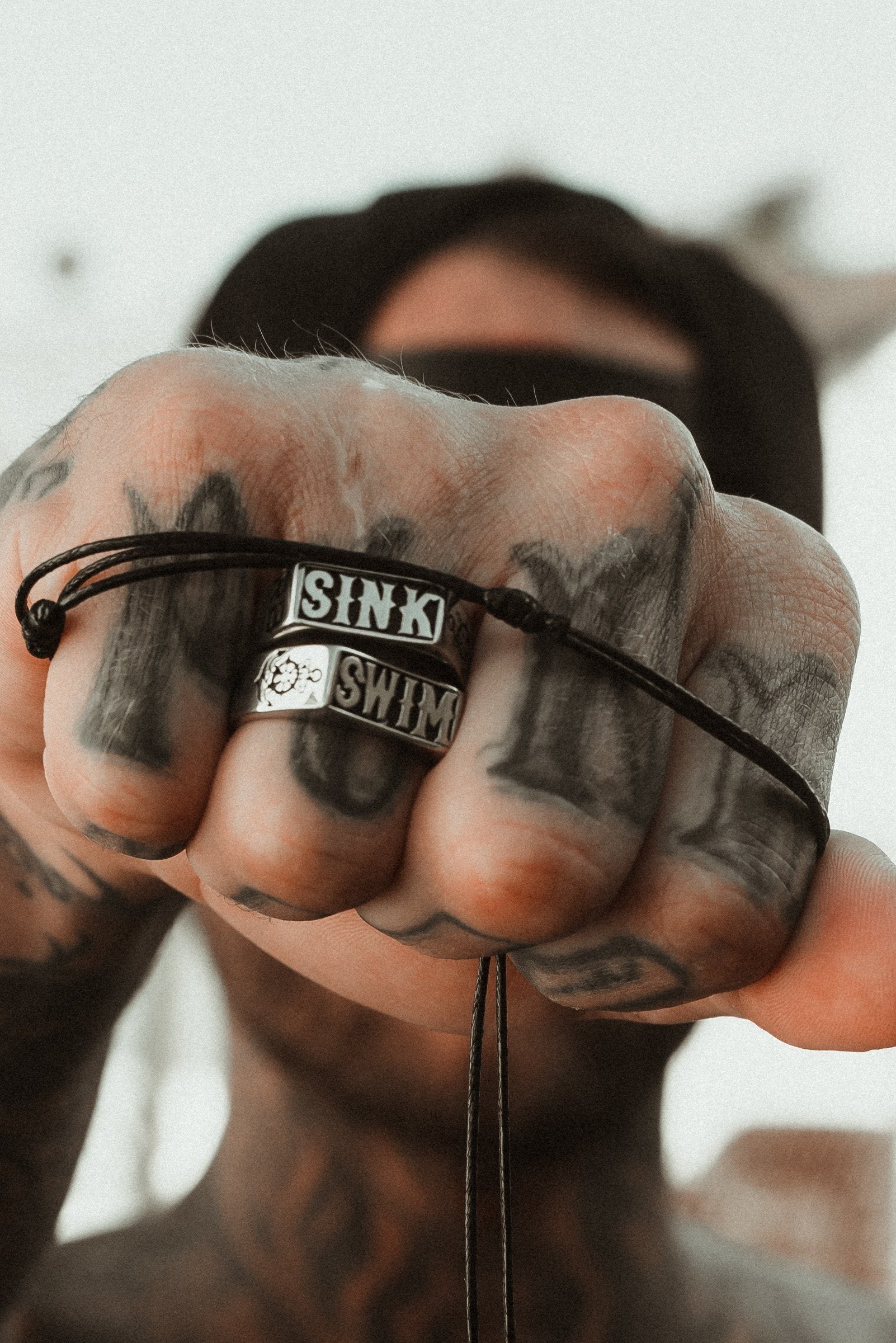 A close-up of a tattooed hand showcases Sink or Swim Rings by Relentless Betrayal on the index and middle fingers, each engraved with SINK and SWIM. A delicate black string wraps loosely around the fingers against a blurred backdrop.