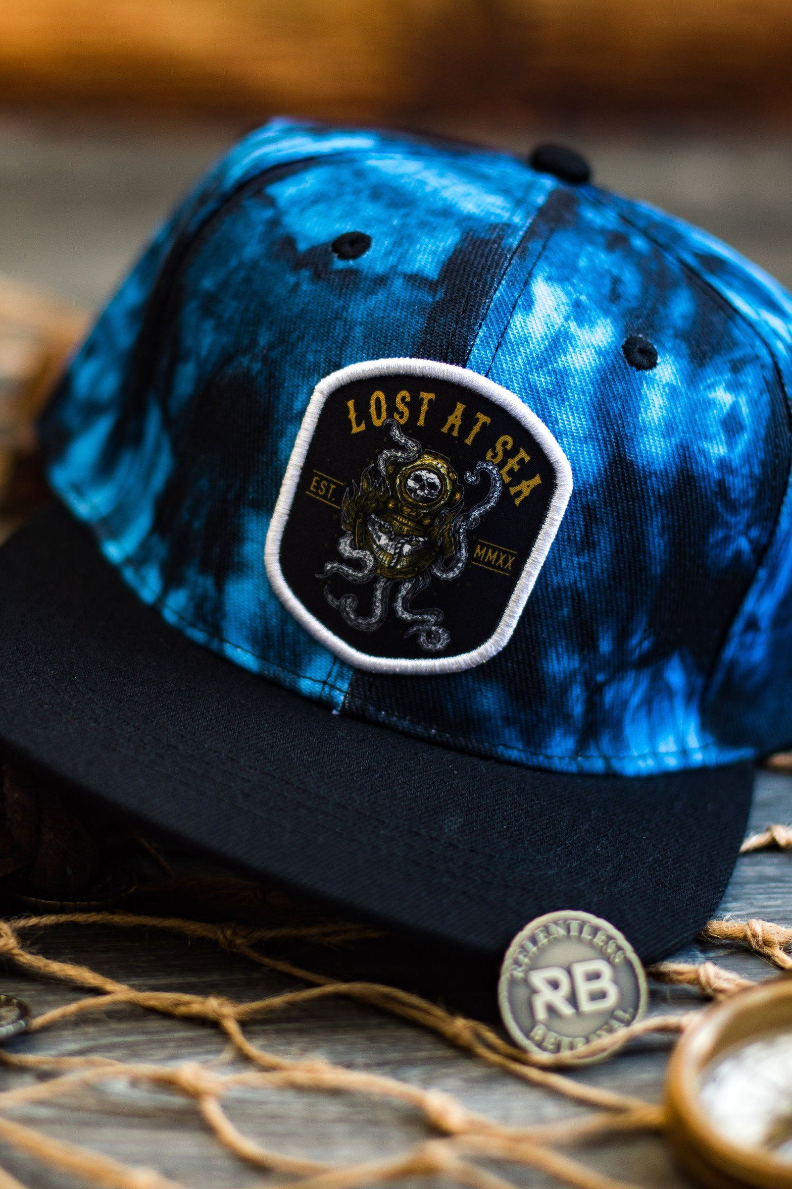 The handmade Lost At Sea Snapback - Tie Dye by Relentless Betrayal showcases a black and blue pattern, a sea creature patch with Lost at Sea text, and a metal RB button on the brim. Its displayed on wood with netting in the background.