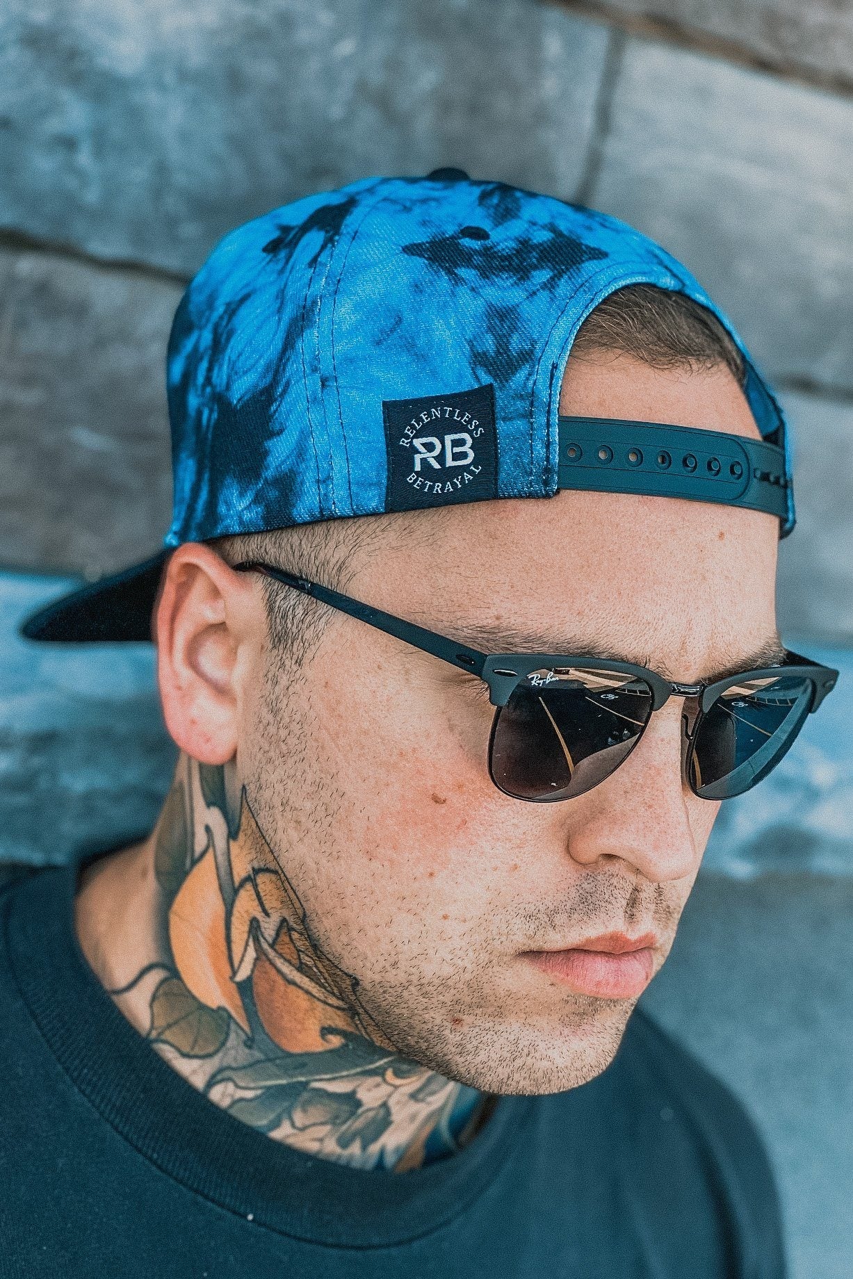A man wears a Lost At Sea Snapback - Tie Dye by Relentless Betrayal backwards with sunglasses. A tattoo adorns his neck above a black shirt, against stone-textured wall panels.