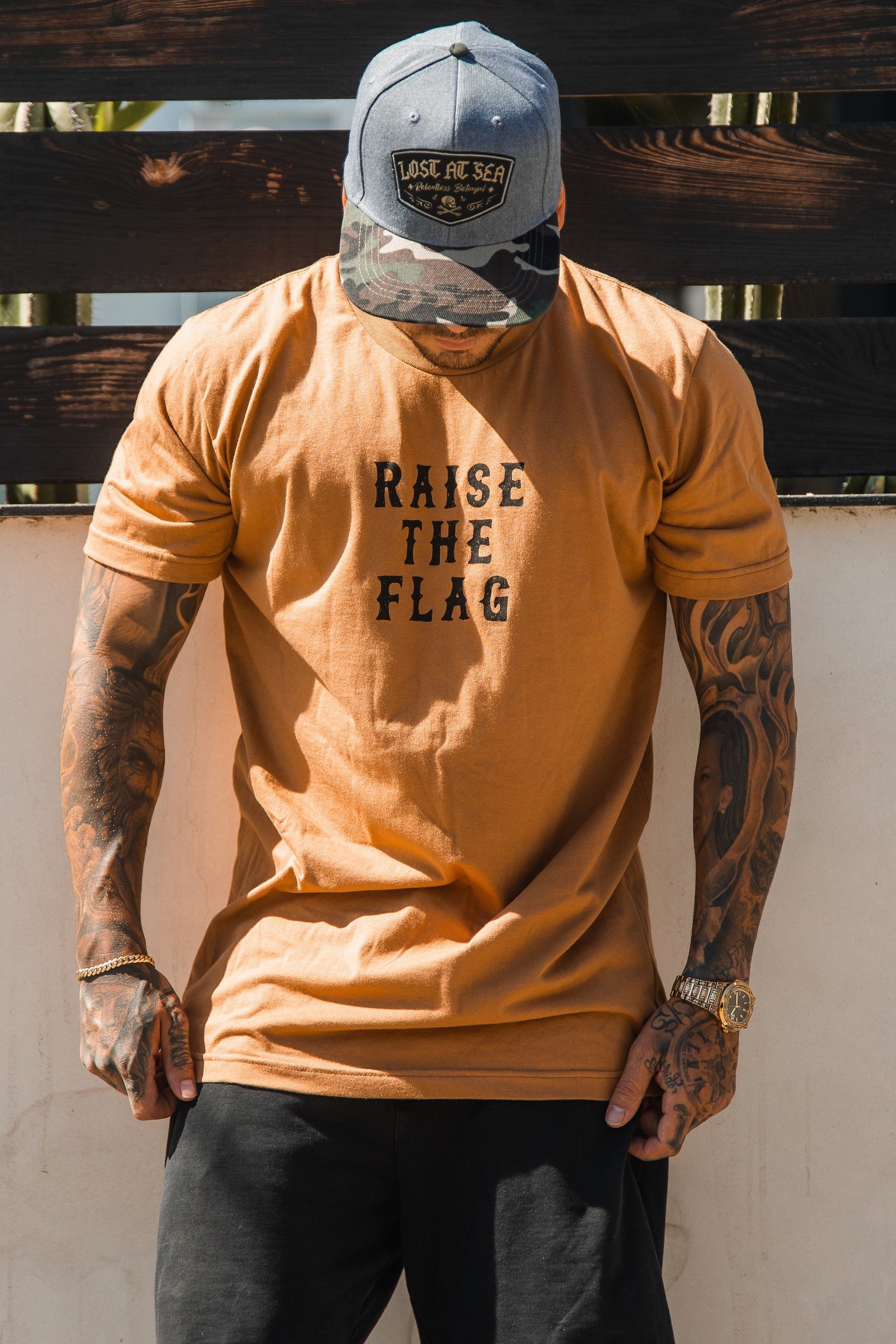 Outdoors, a person stands wearing a tan Raise the Flag Tee by Relentless Betrayal, made of soft jersey cotton. They pair it with a camo cap and dark pants. Tattooed arms reminiscent of a pirate flag hang by their sides.