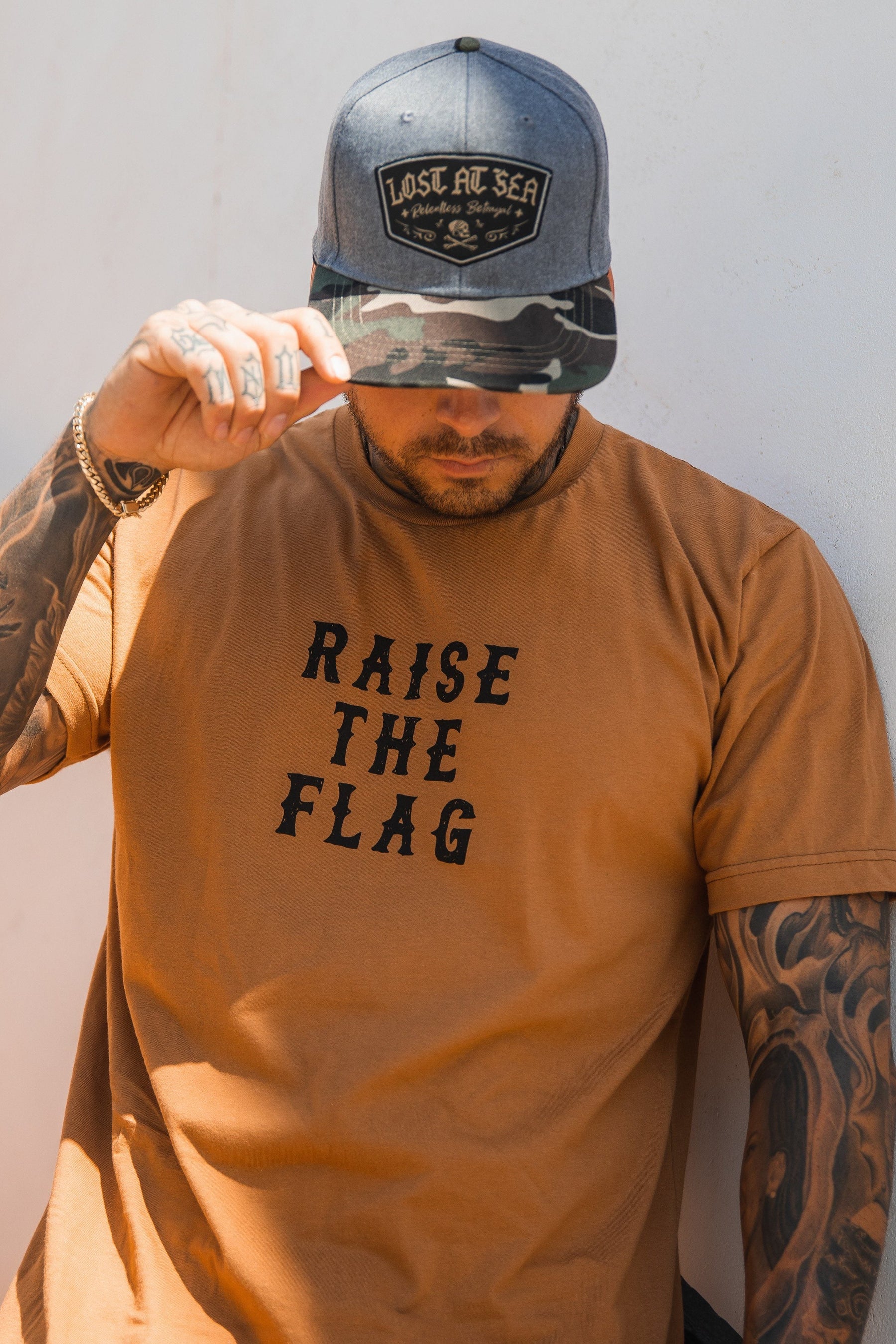 A tattooed individual wears a brown Raise the Flag Tee by Relentless Betrayal and a cap with a LOST AT SEA patch. The tilted cap channels pirate vibes as they pose against a white wall.