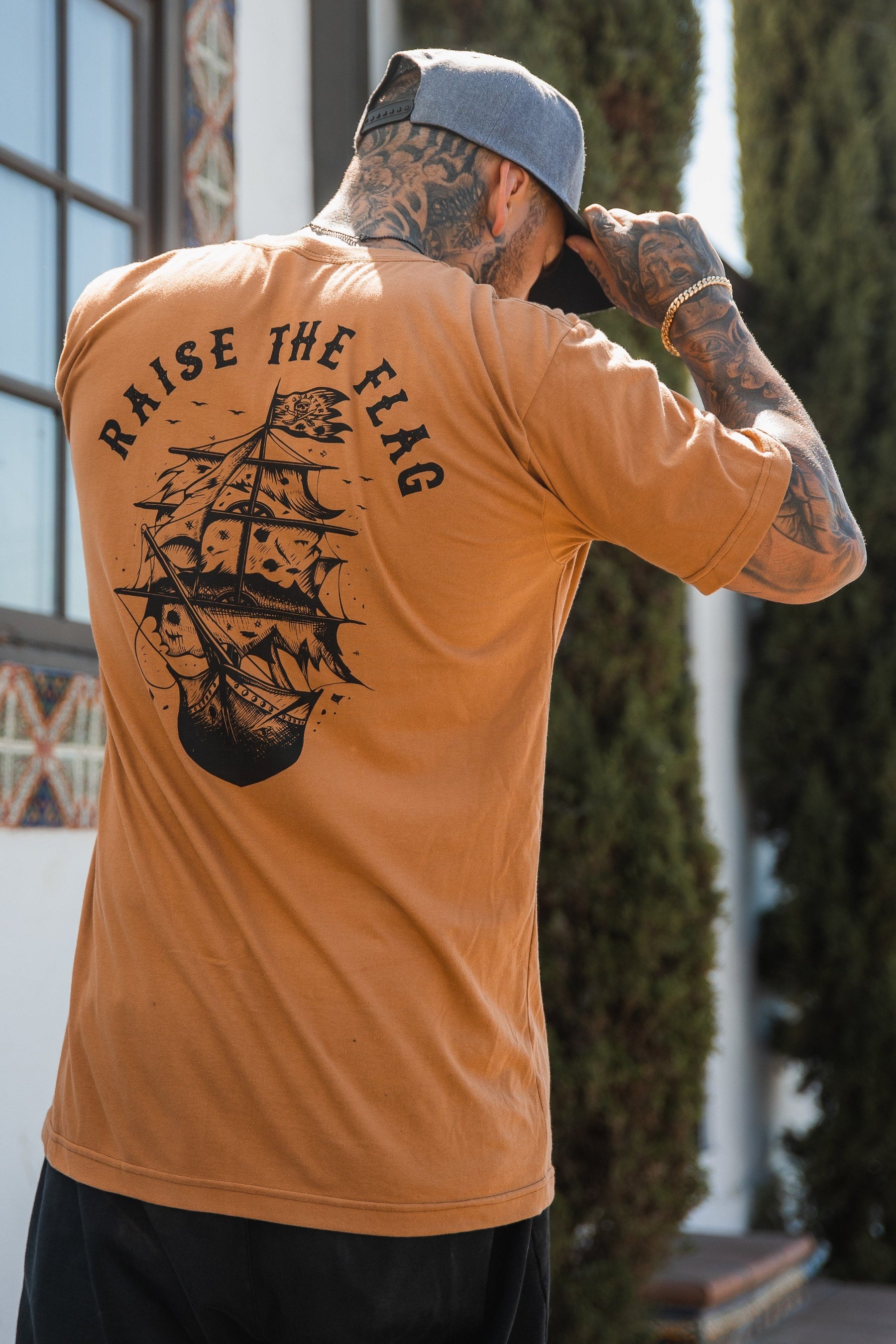 Near a tree and building, a tattooed individual adjusts their cap while wearing the Relentless Betrayals Raise the Flag Tee in brown jersey cotton. The T-shirt, adorned with a pirate ship and RAISE THE FLAG text on the back, captures a daring spirit of surrender or face an attack.