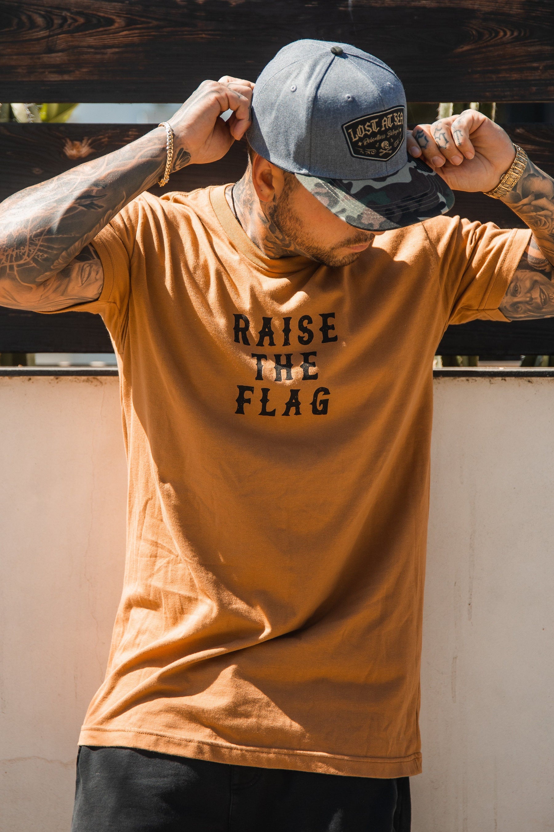 A tattooed man in a Relentless Betrayal Raise the Flag Tee adjusts his cap against a light-paneled backdrop. His downward-tilted head obscures his face.