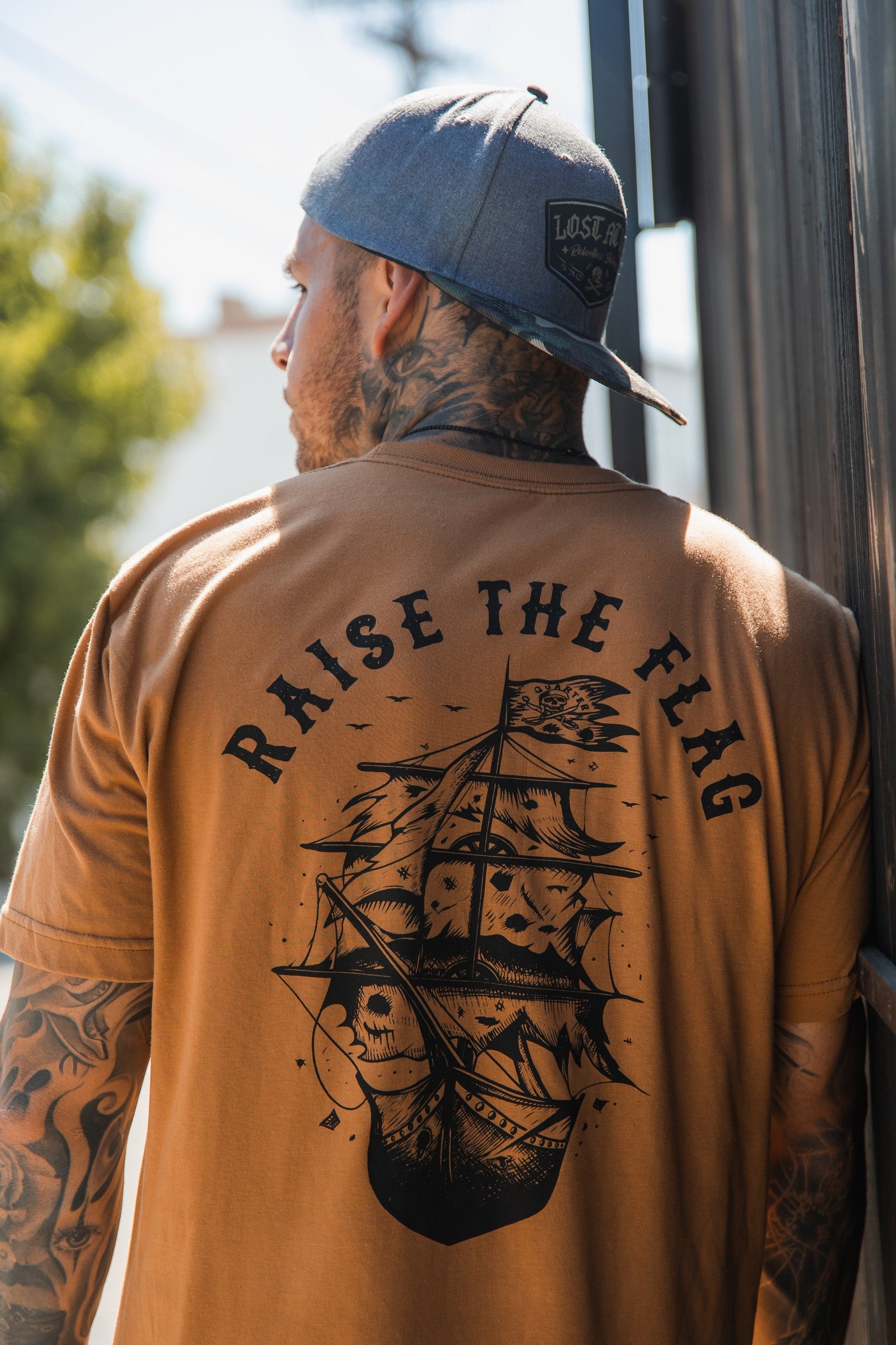 Outdoors by a building, a man in the Relentless Betrayals Raise the Flag Tee with ship and RAISE THE FLAG design, flaunts tattoos on his arms and neck. A backward baseball cap completes his look, evoking the bold spirit of an unfurled pirate flag.
