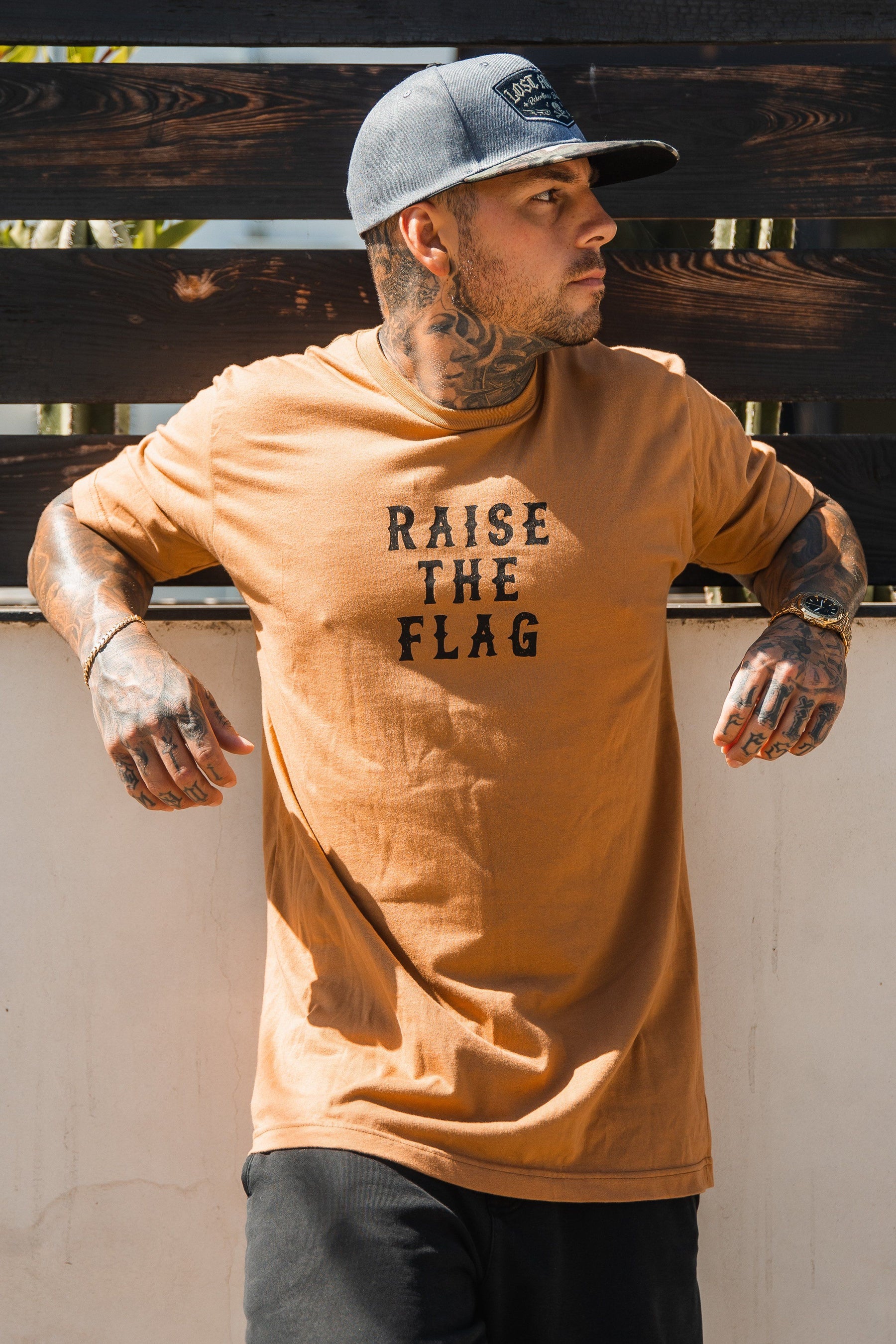 A tattooed man leans against a white wall in a tan Raise the Flag Tee by Relentless Betrayal, paired with black pants and a gray cap. He gazes right as wooden planks and lush greenery evoke pirate vibes in the background.