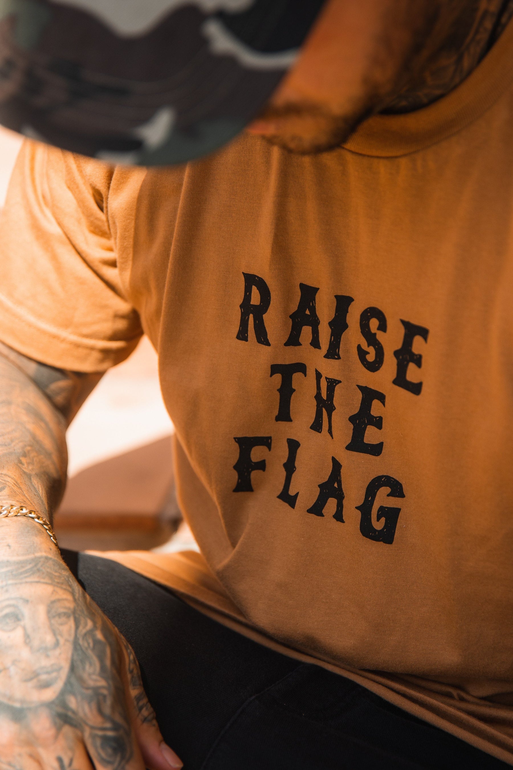 A person dons a Relentless Betrayal Raise the Flag Tee, reminiscent of a pirate flag, paired with a camouflage cap. Tattoos accentuate their arm, enhancing the rebellious look, with the focus on their upper body and striking shirt.