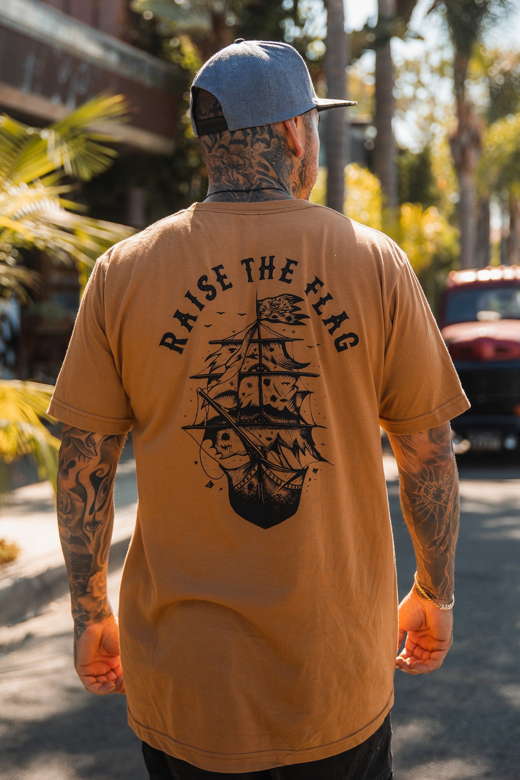 A bearded tattooed person wearing a cap stands on a sunny street in a brown Raise the Flag Tee from Relentless Betrayal, featuring a ship and RAISE THE FLAG on the back. Swaying palm trees and a red truck enhance the laid-back vibe of the scene.