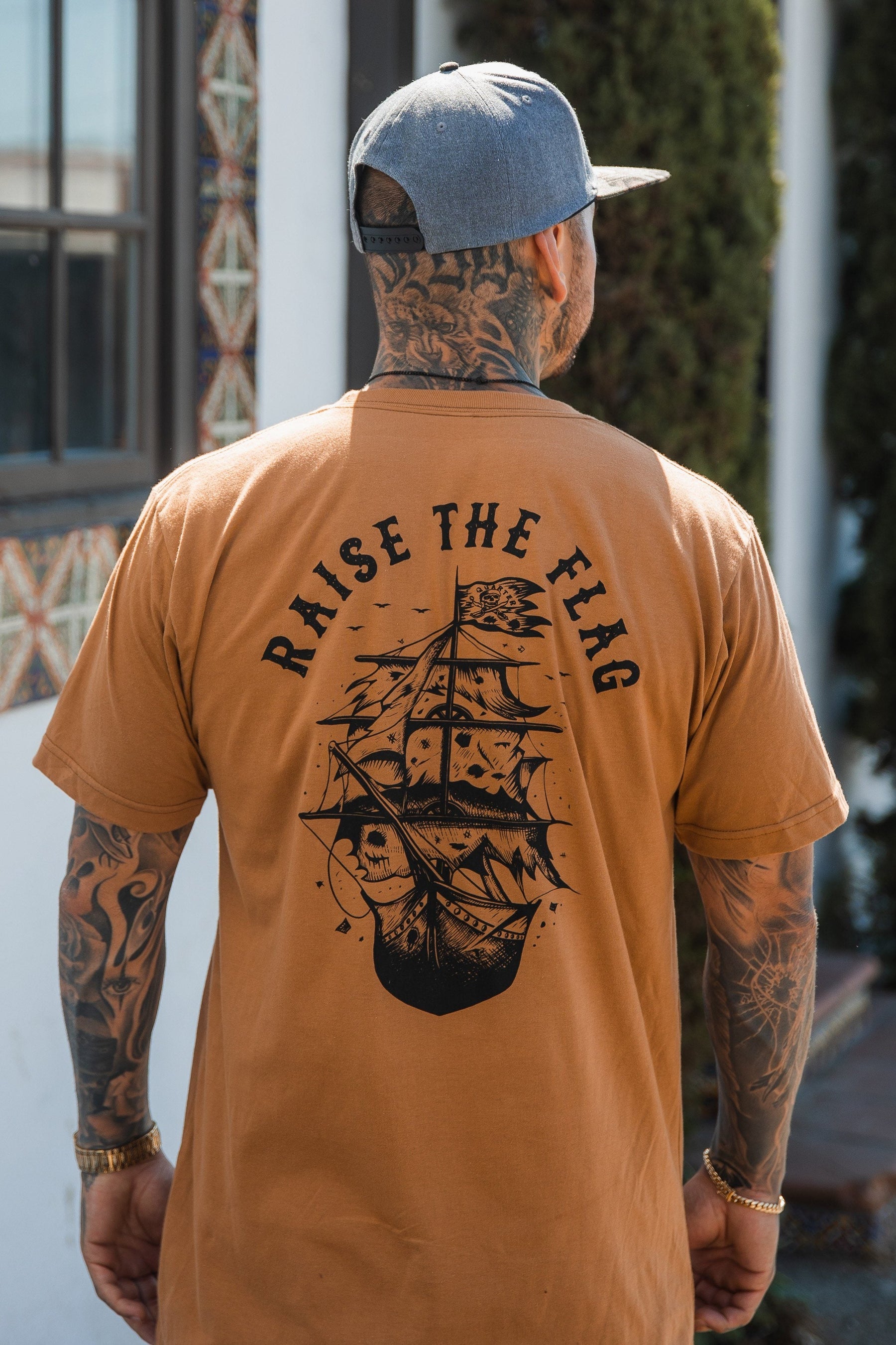 A tattooed person wears Relentless Betrayals Raise the Flag Tee, a brown jersey cotton shirt with a ship graphic and RAISE THE FLAG on the back. Standing outdoors by a textured wall and large trees, their gray cap embodies an untamed pirate spirit.