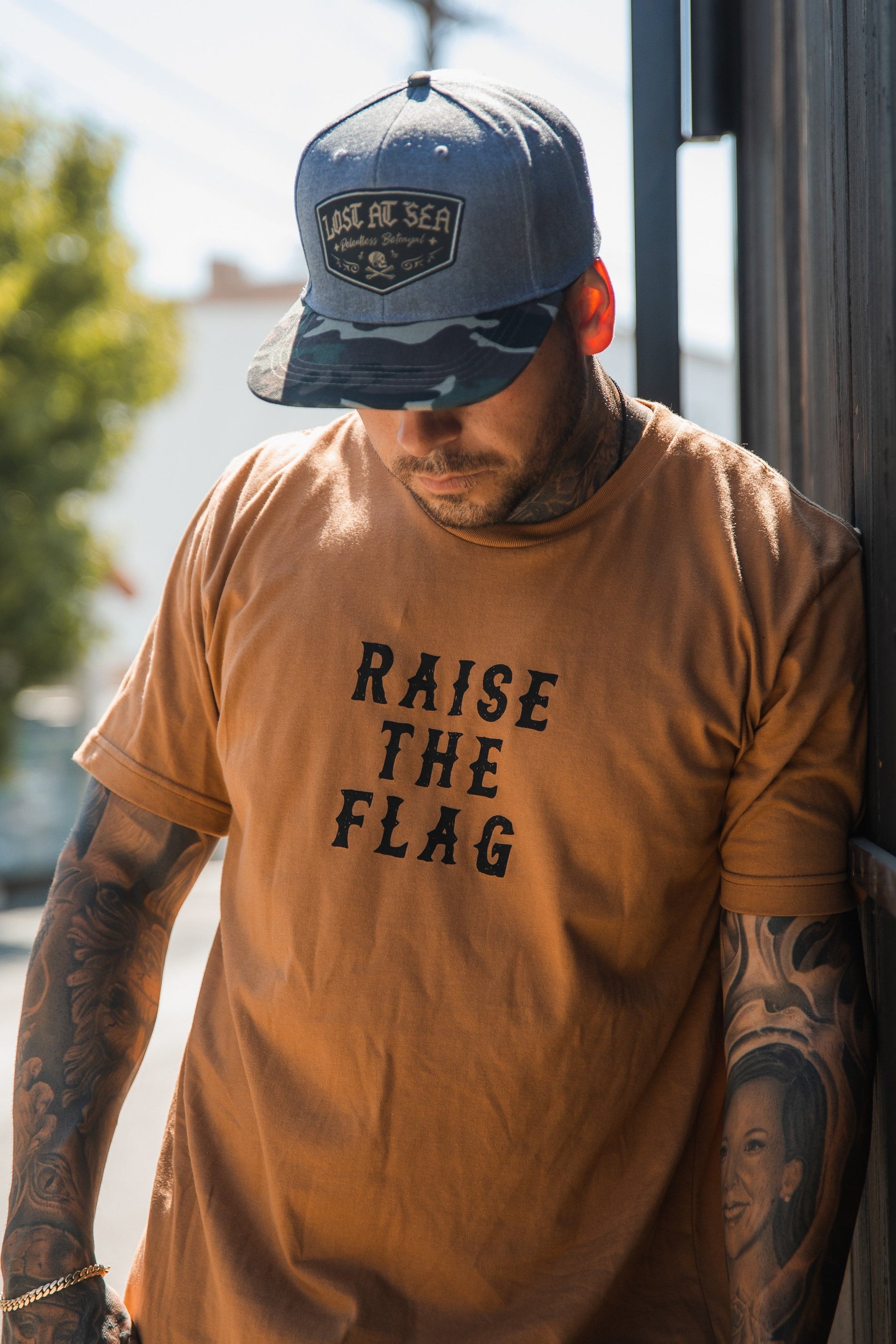 A tattooed man stands outdoors, wearing Relentless Betrayals Raise the Flag Tee and a gray Lost At Sea cap. The sunlight creates shadows around him, reminiscent of a pirate flag in the wind.