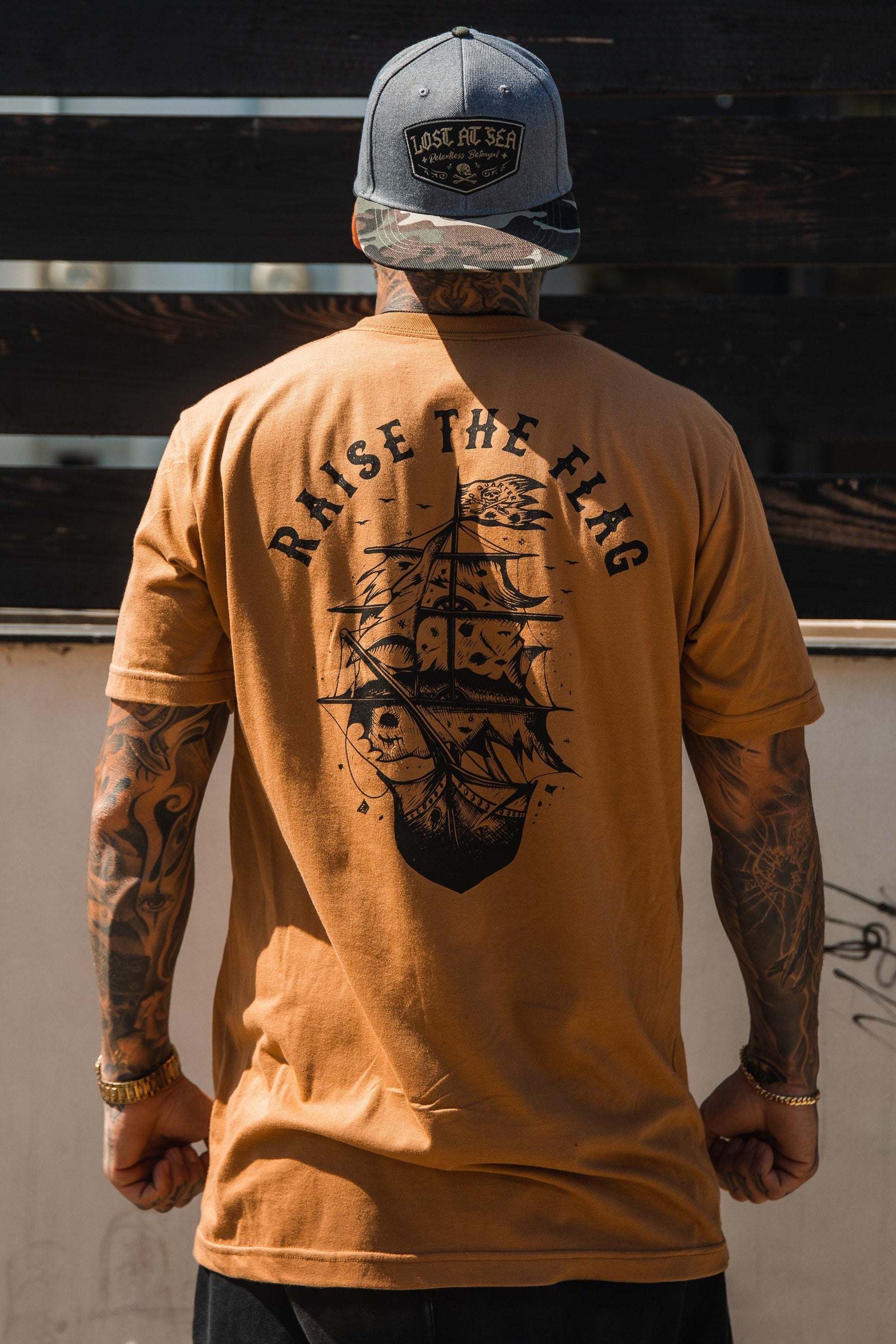 With their back to the camera, a person wears the Raise the Flag Tee by Relentless Betrayal, featuring a ship design and RAISE THE FLAG. Tattoos cover their skin, and a camo baseball cap heightens the rebellious pirate flag vibe.