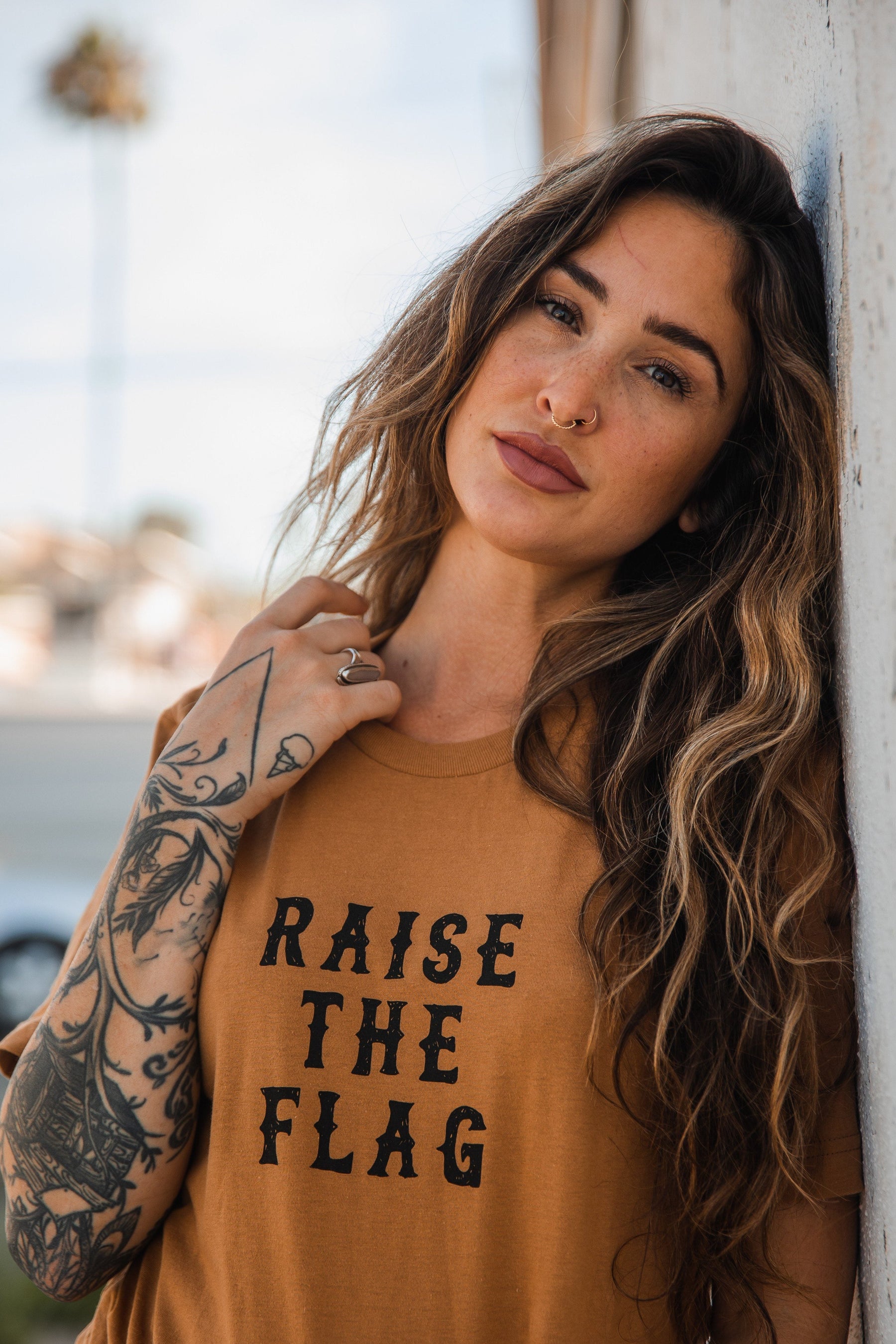 A person with long, wavy hair and tattoos leans against a wall, wearing the Raise the Flag Tee by Relentless Betrayal. The shirt features RAISE THE FLAG, evoking a pirate flag, while the blurred background suggests an outdoor setting.