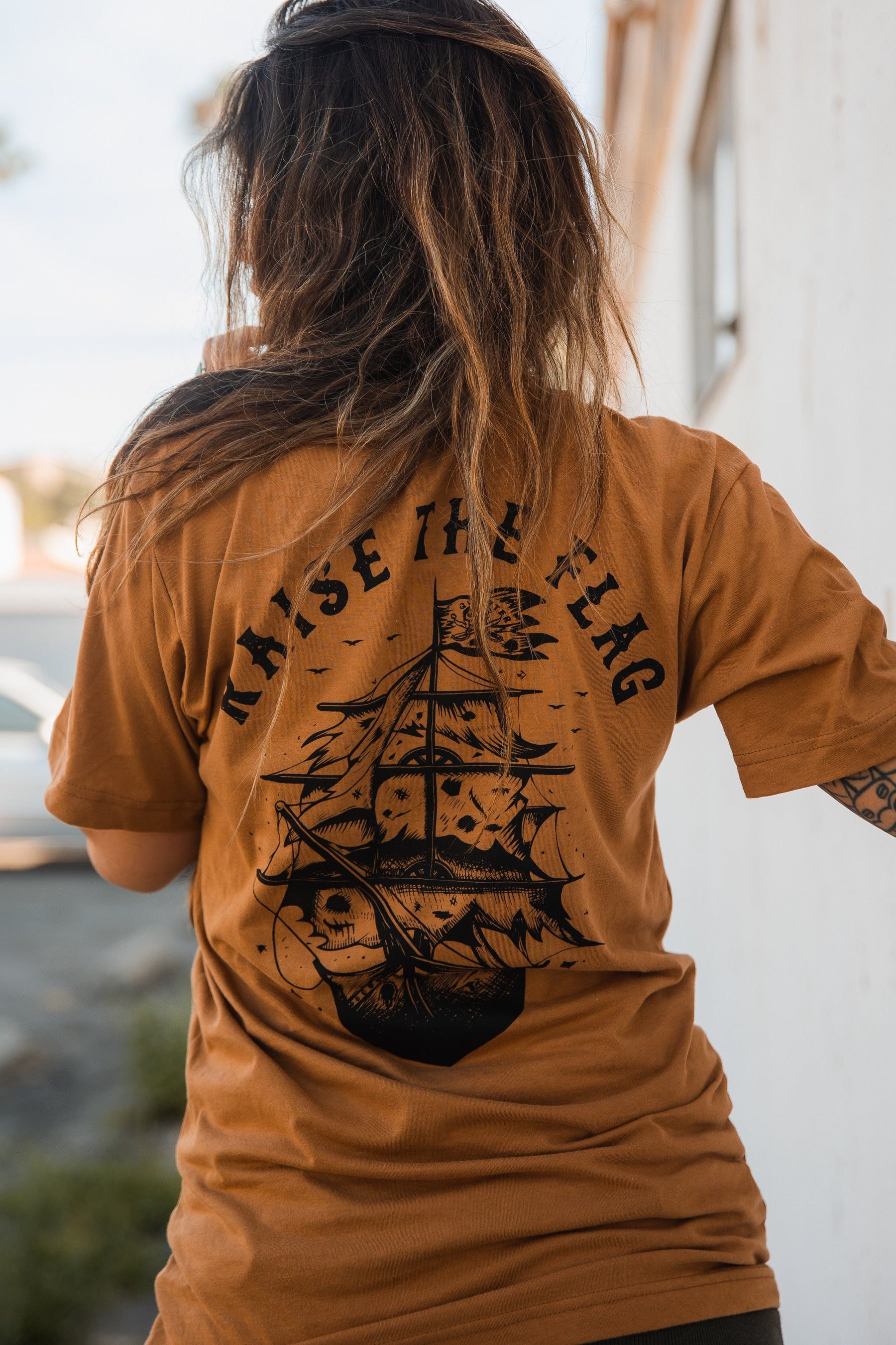 Facing away from a light-colored wall outdoors, a person with long hair wears the Relentless Betrayals Raise the Flag Tee, a brown jersey cotton T-shirt featuring a bold black sailing ship design and the slogan RAISE THE FLAG on the back, evoking a pirate flag.