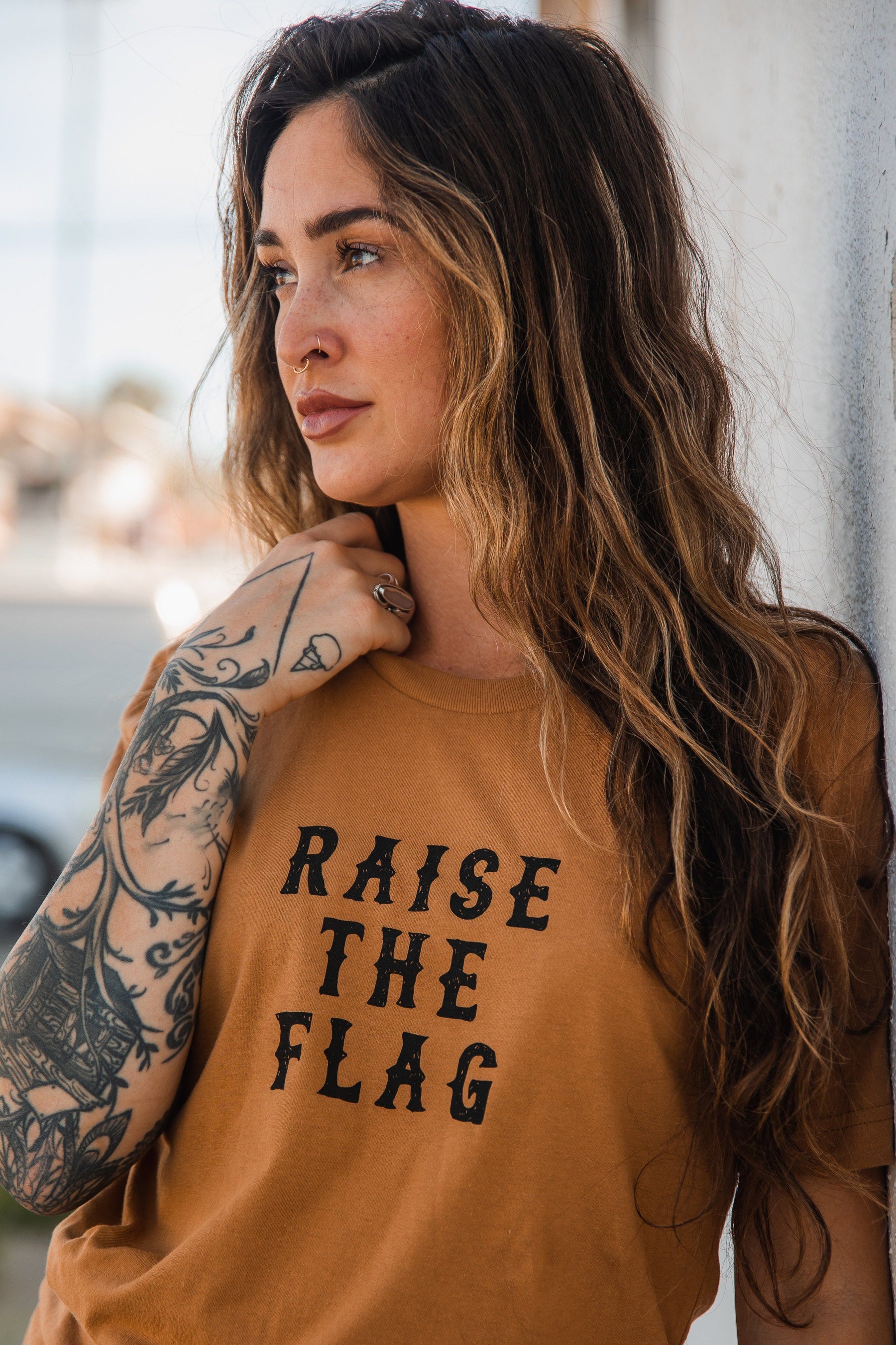 A woman with long wavy hair and tattoos on her right arm leans against a wall, wearing a brown Raise the Flag Tee by Relentless Betrayal. Her pensive expression is turned left against a softly blurred backdrop.